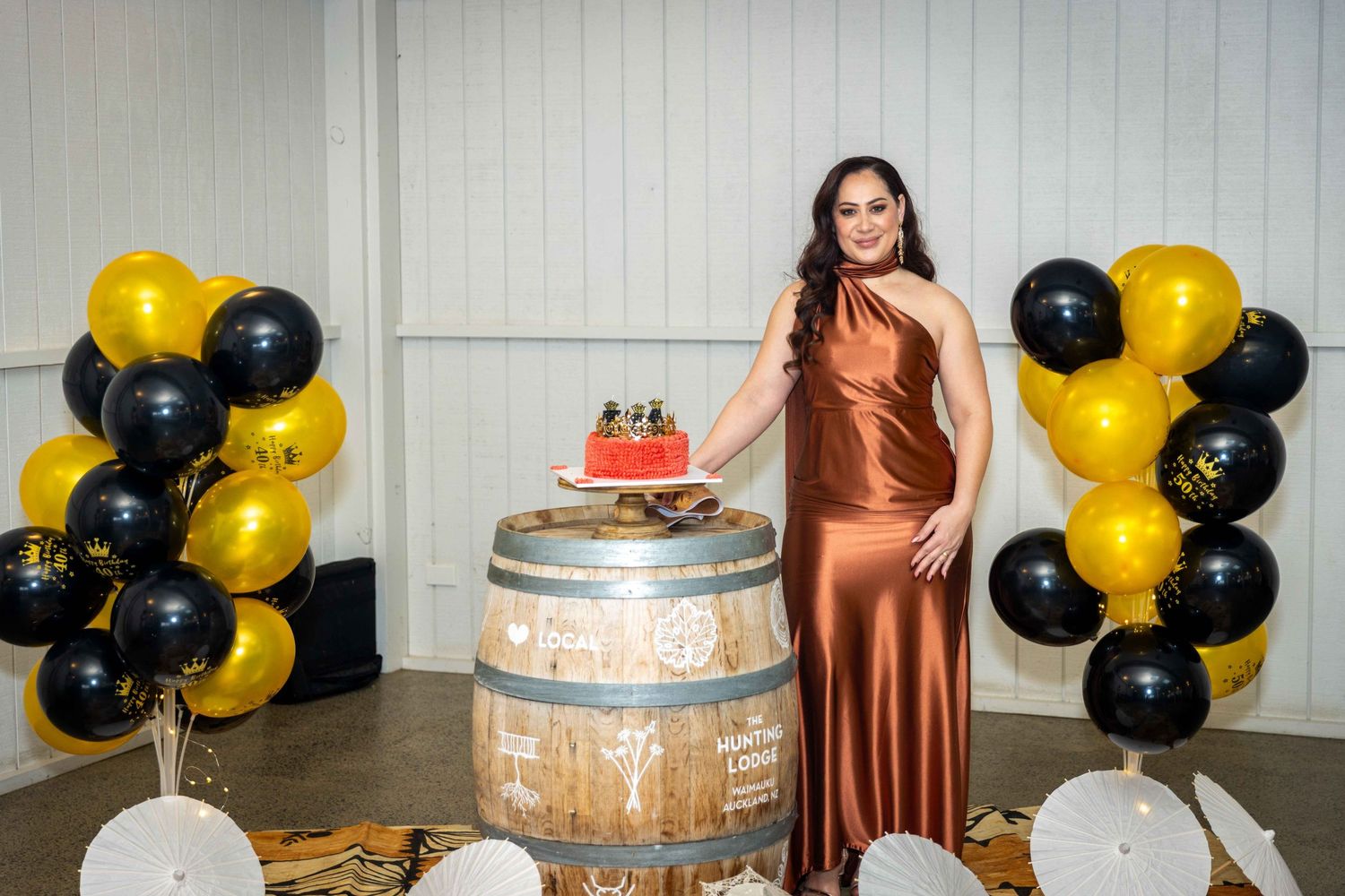 Woman in copper satin gown posing beside birthday cake on barrel with black and gold balloons.