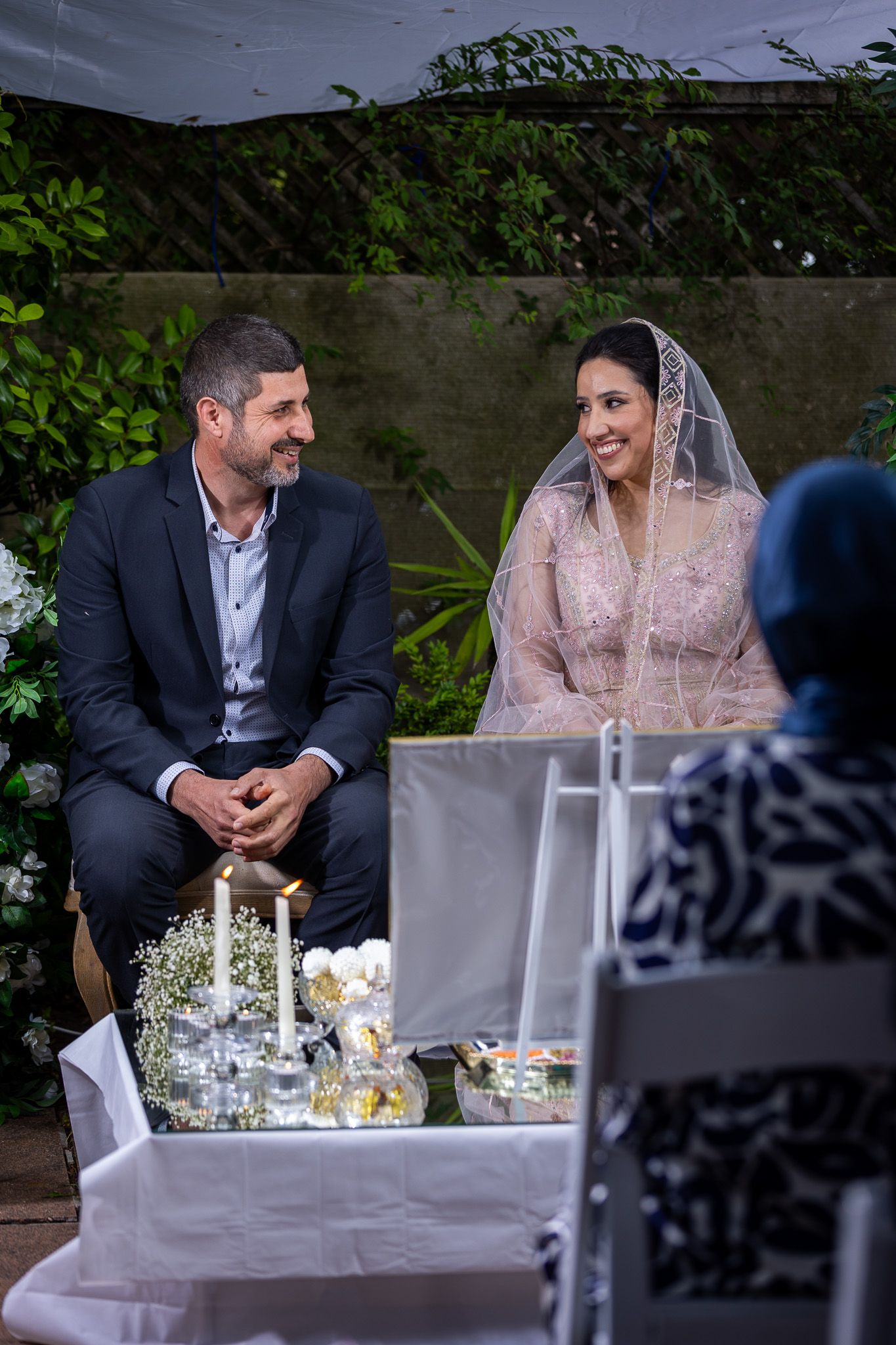 Couple smiling at their wedding ceremony, bride in lace veil and groom in suit, seated at decorated table outdoors.