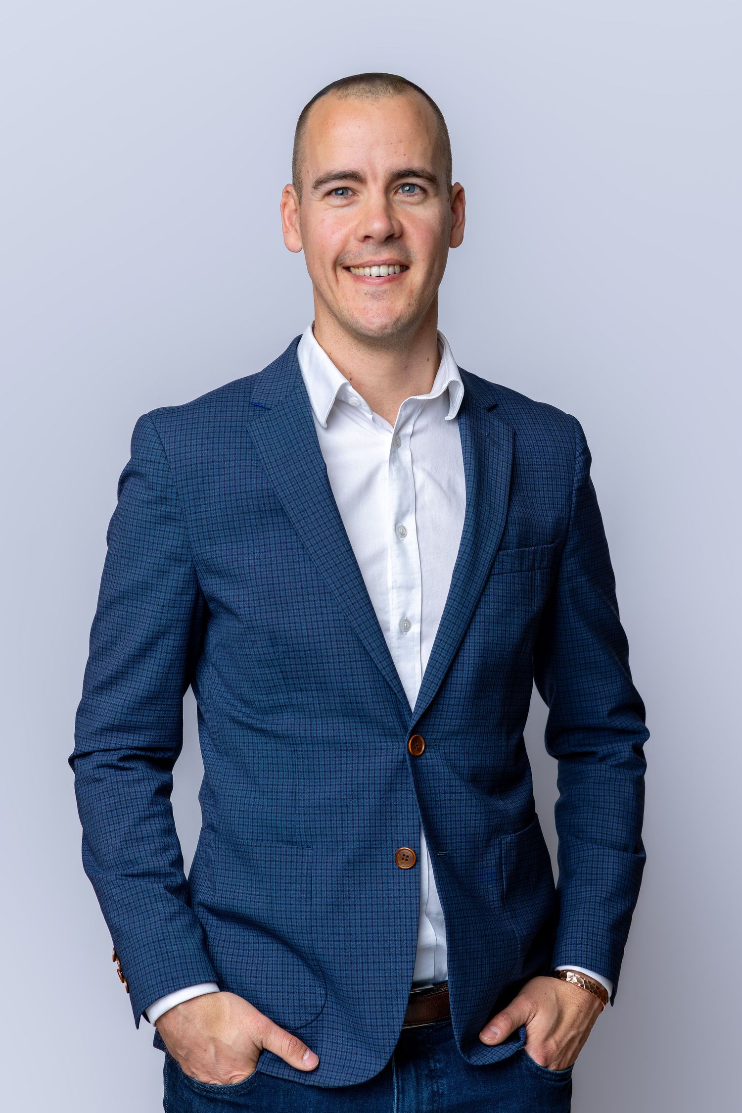 Smiling man in blue checkered blazer and white shirt poses confidently with hands in pockets against gray background.