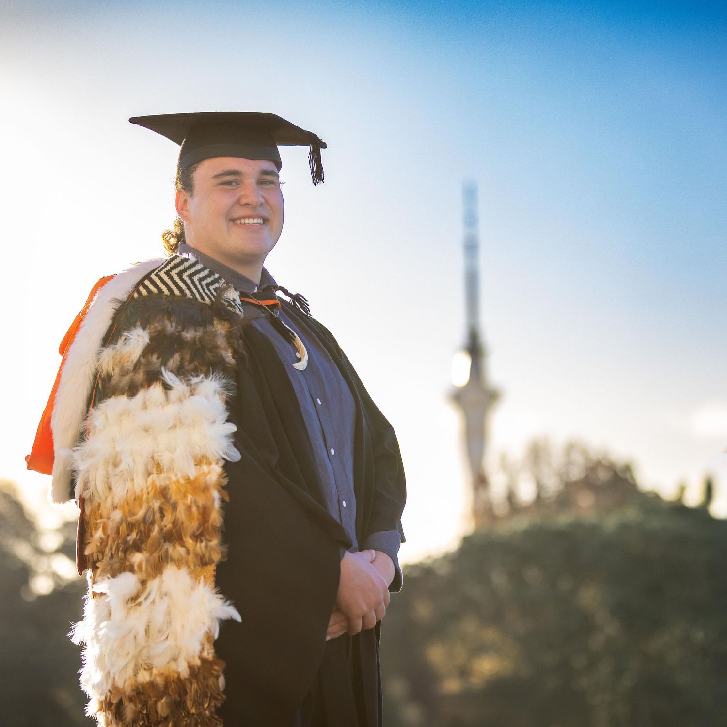 Graduate in black cap and gown with traditional Maori cloak smiles outdoors with blue sky and tower behind.
