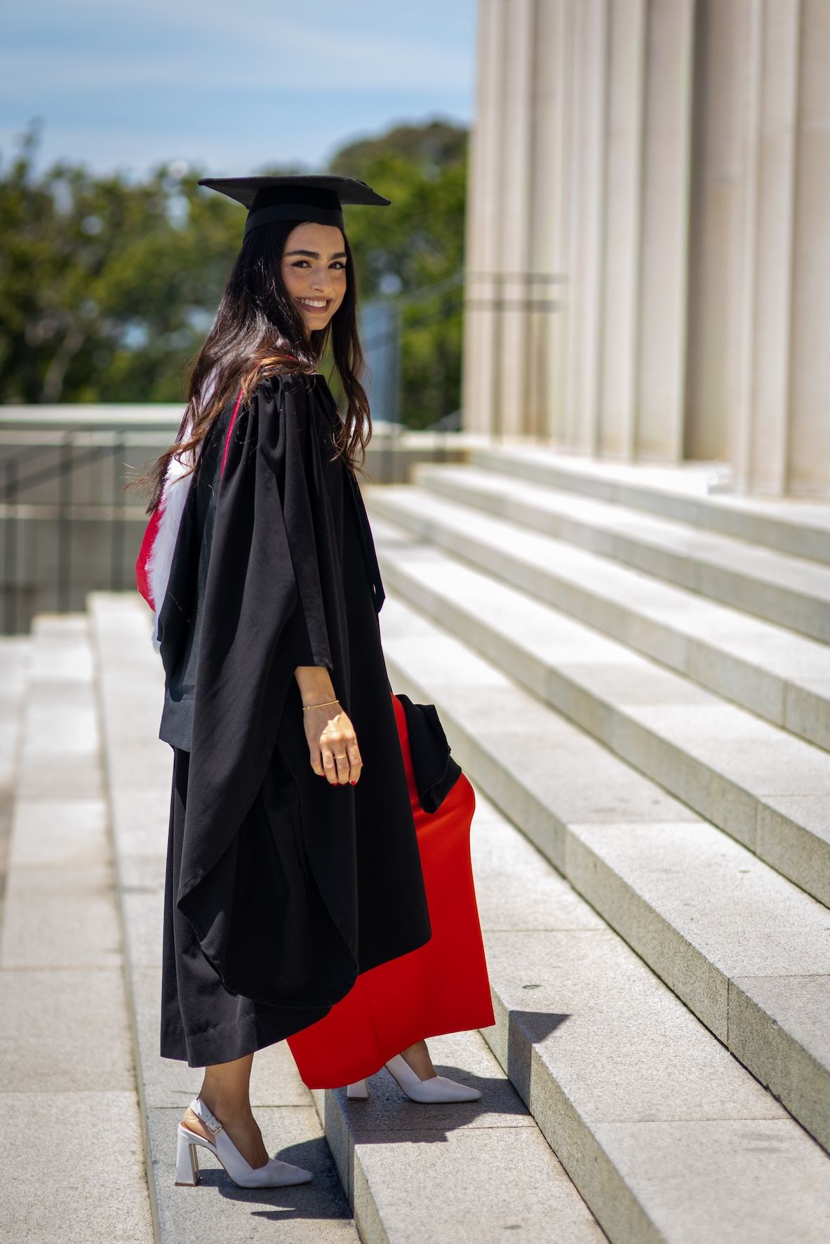 Young woman in black graduation cap and gown with red accents smiles on university steps on a sunny day.