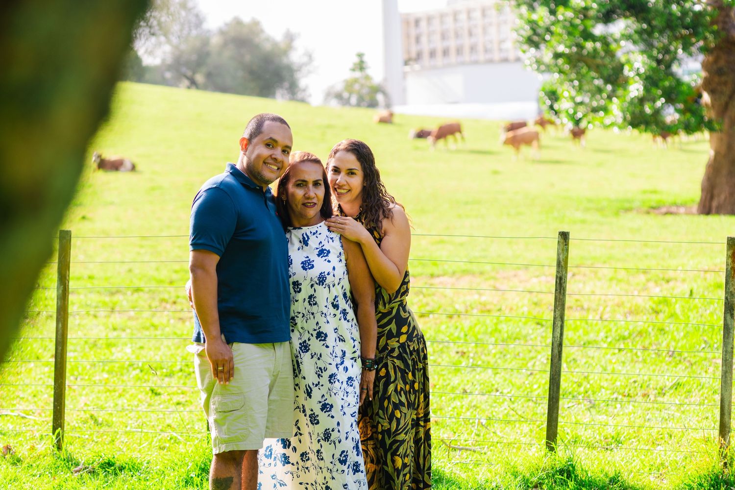 Three people smiling together on a green farm with grazing cows and a building in the background.