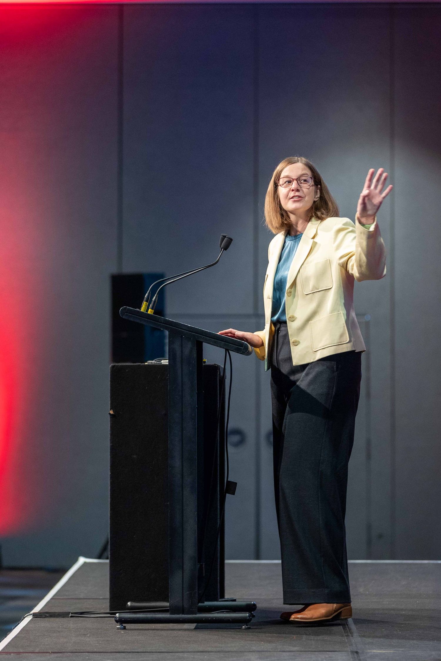 A speaker in a yellow blazer gestures while presenting at a podium on stage with dramatic lighting.