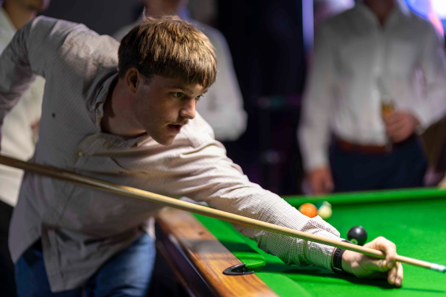 Young male snooker player leaning over green baize table, taking a precise shot with cue in a dimly lit billiards hall.