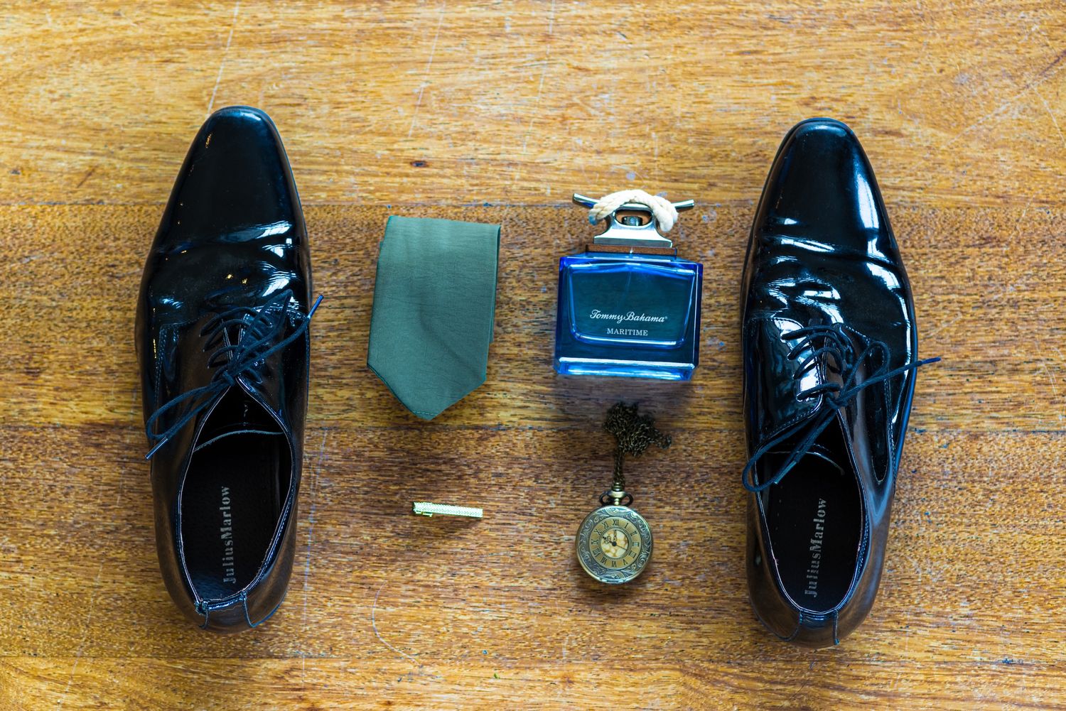 Black patent leather dress shoes, green tie, cologne, cufflinks, and pocket watch arranged on wooden surface.