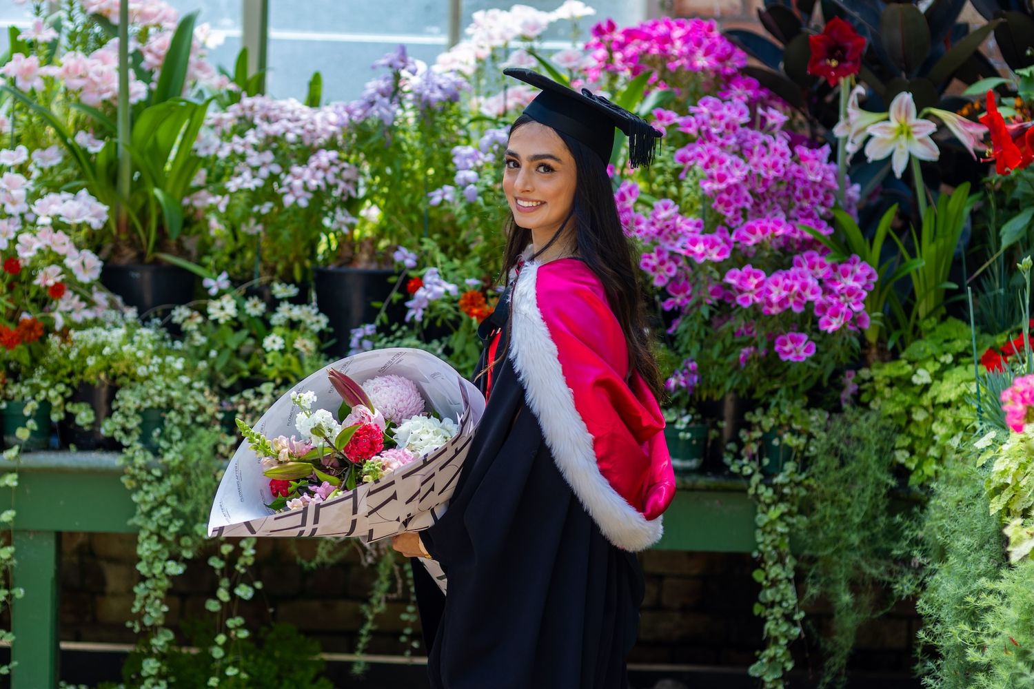 Woman in graduation cap and gown holding flower basket surrounded by colorful blooms in lush garden setting.