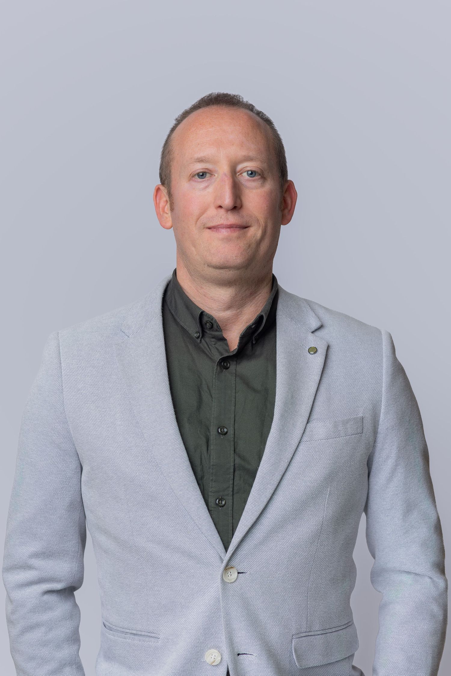 Professional man in light gray blazer and dark green shirt poses confidently against a neutral gray background.