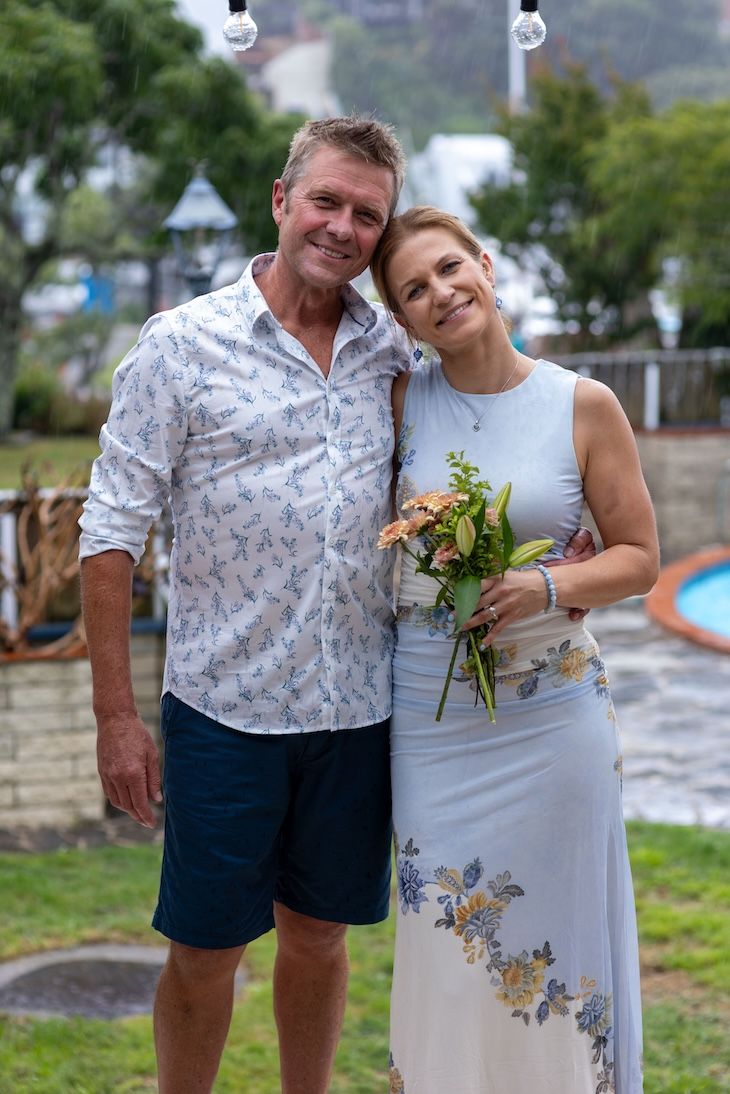 Happy couple posing outdoors, woman holding bouquet, man in floral shirt, casual wedding attire, garden background.