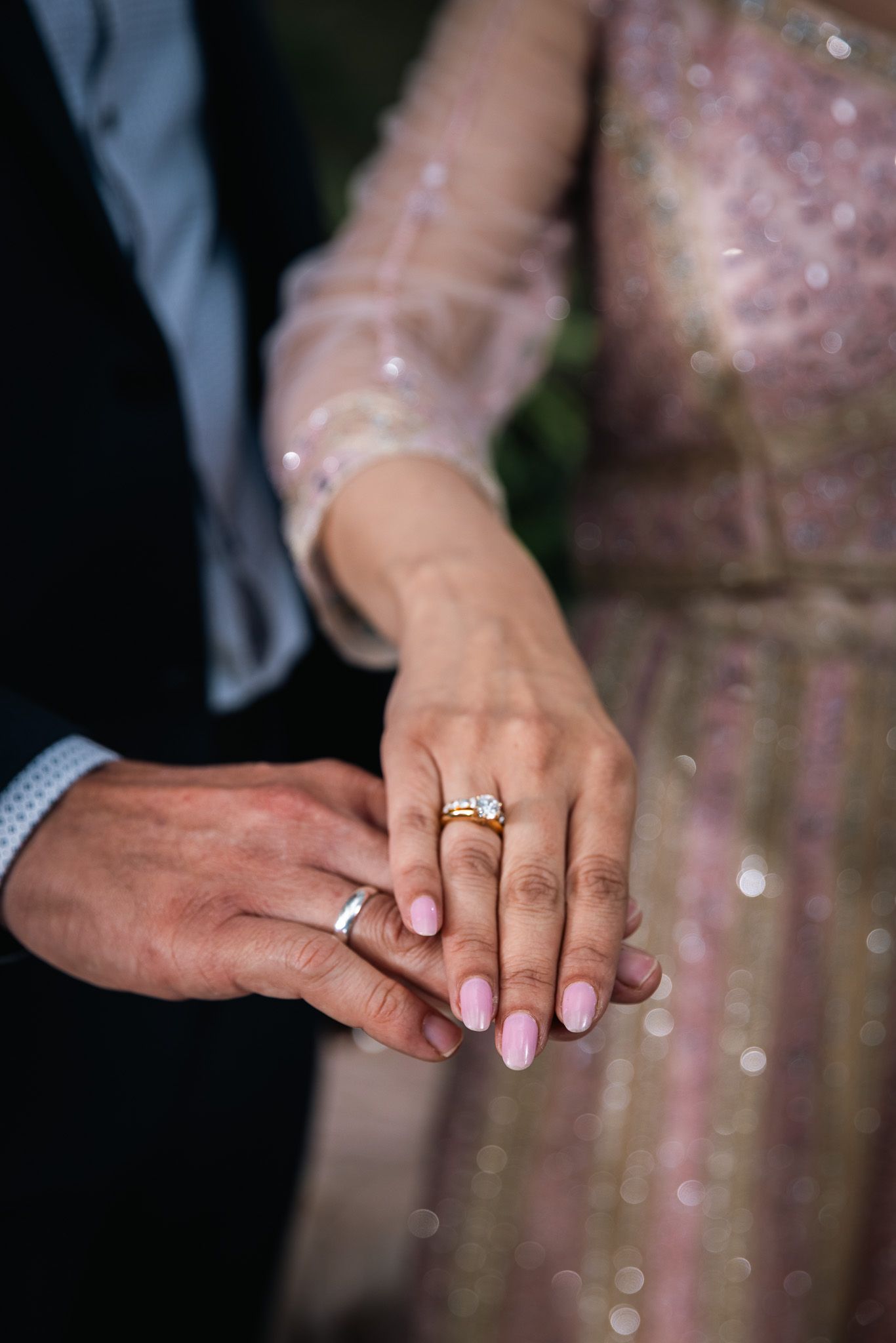 Couple holding hands showing wedding rings, woman wearing pink sequin dress with manicured nails.