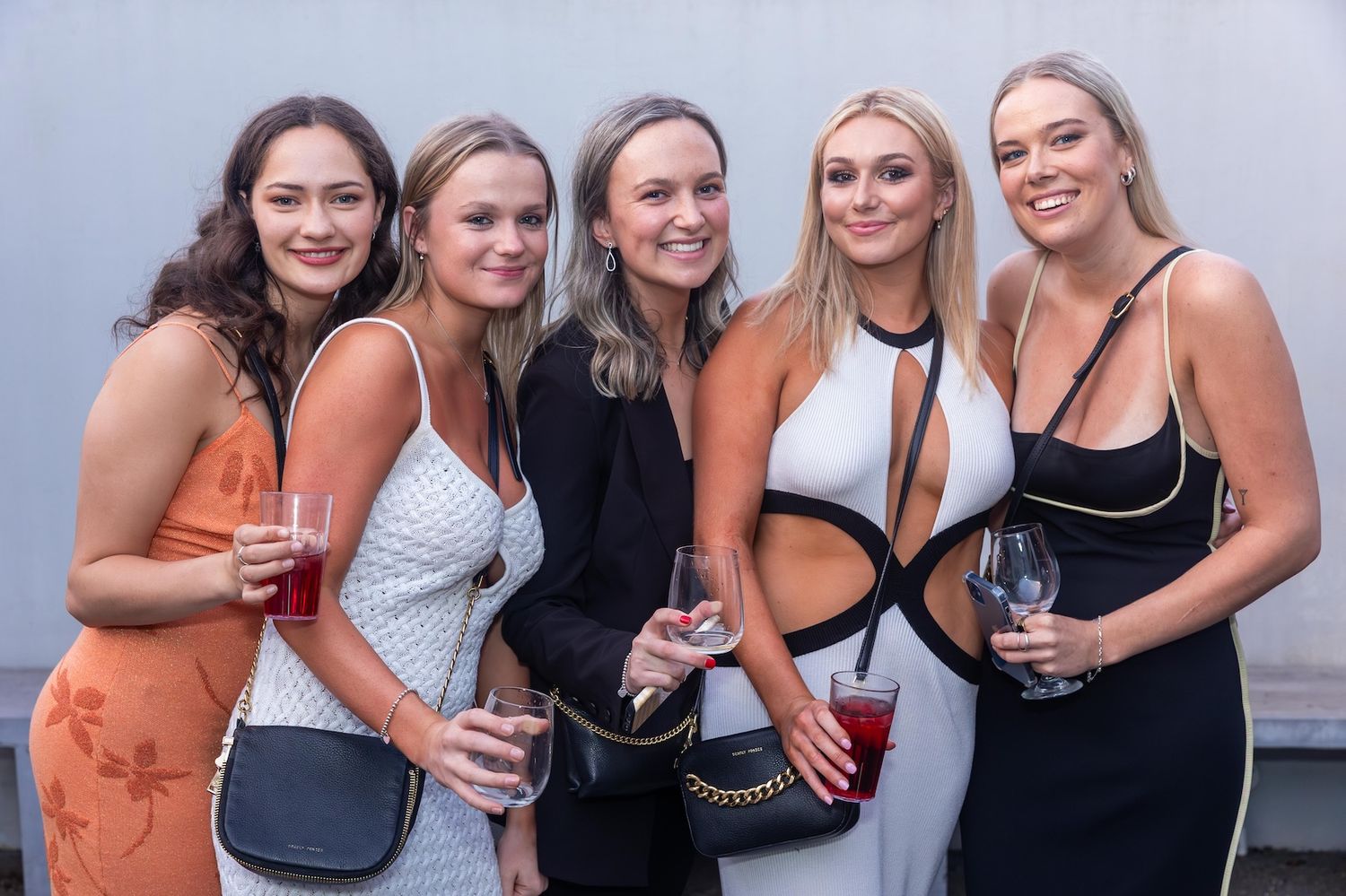 Five young women smiling and holding drinks at an outdoor social event, dressed in elegant evening attire.