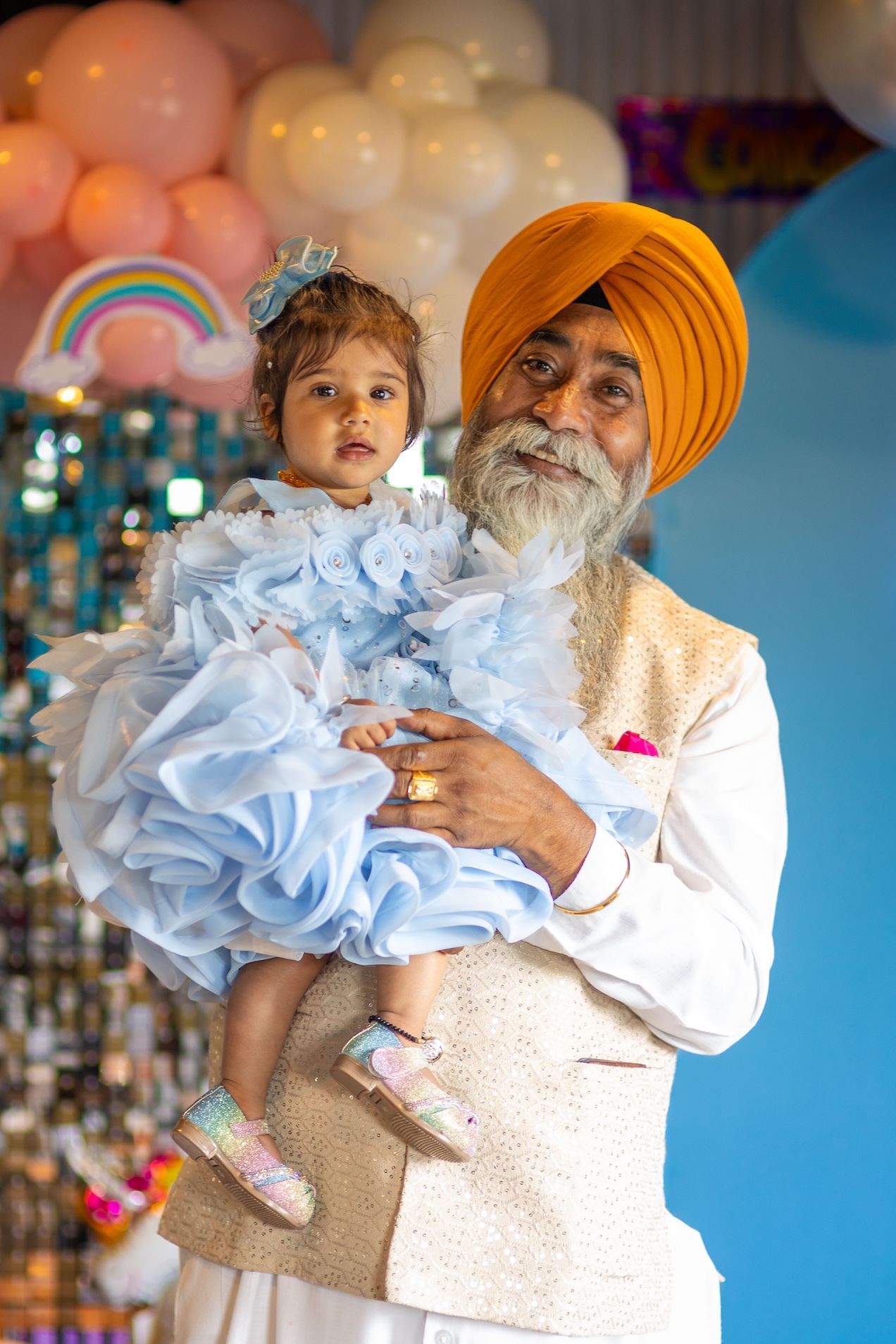 Sikh grandfather in orange turban holds baby girl in blue ruffled dress at a colorful balloon party.
