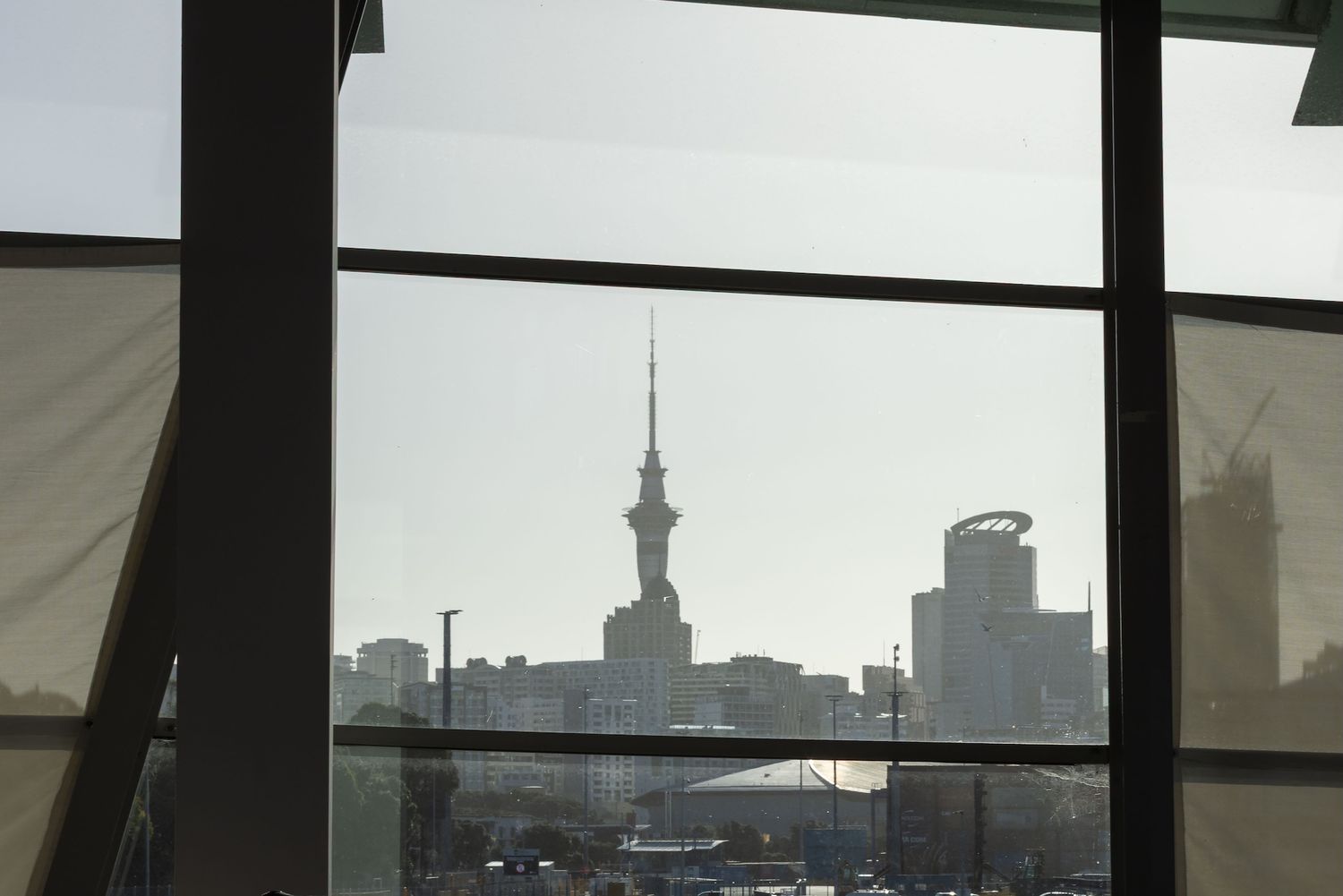 View of Auckland Sky Tower through large windows with city skyline and parking lot visible on a clear day.