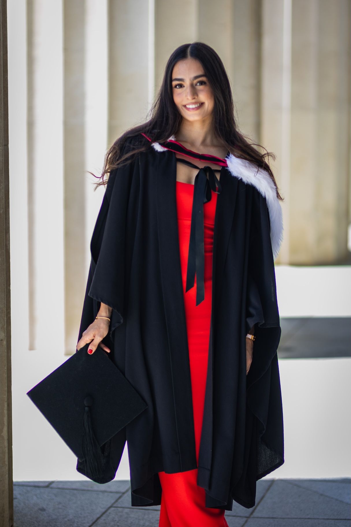 Young woman in black graduation gown with white fur trim and red dress, holding mortarboard cap, smiling outdoors.