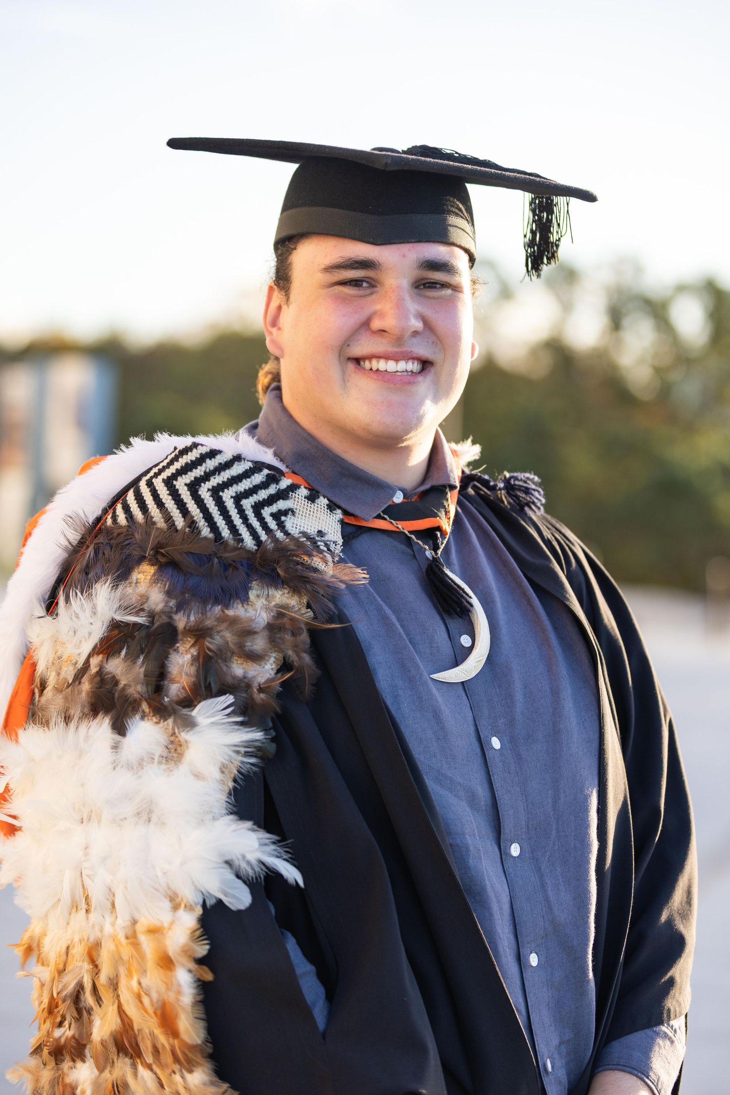 Smiling graduate in cap and gown wearing traditional Maori cloak with feathers and pounamu pendant outdoors.