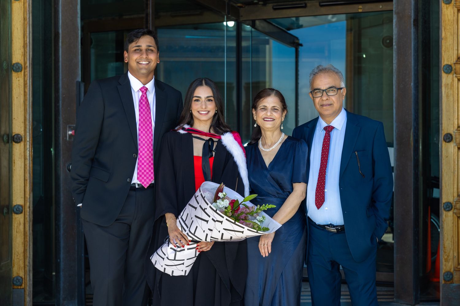 Graduate in cap and gown holds flowers, posing with three family members outside a building.