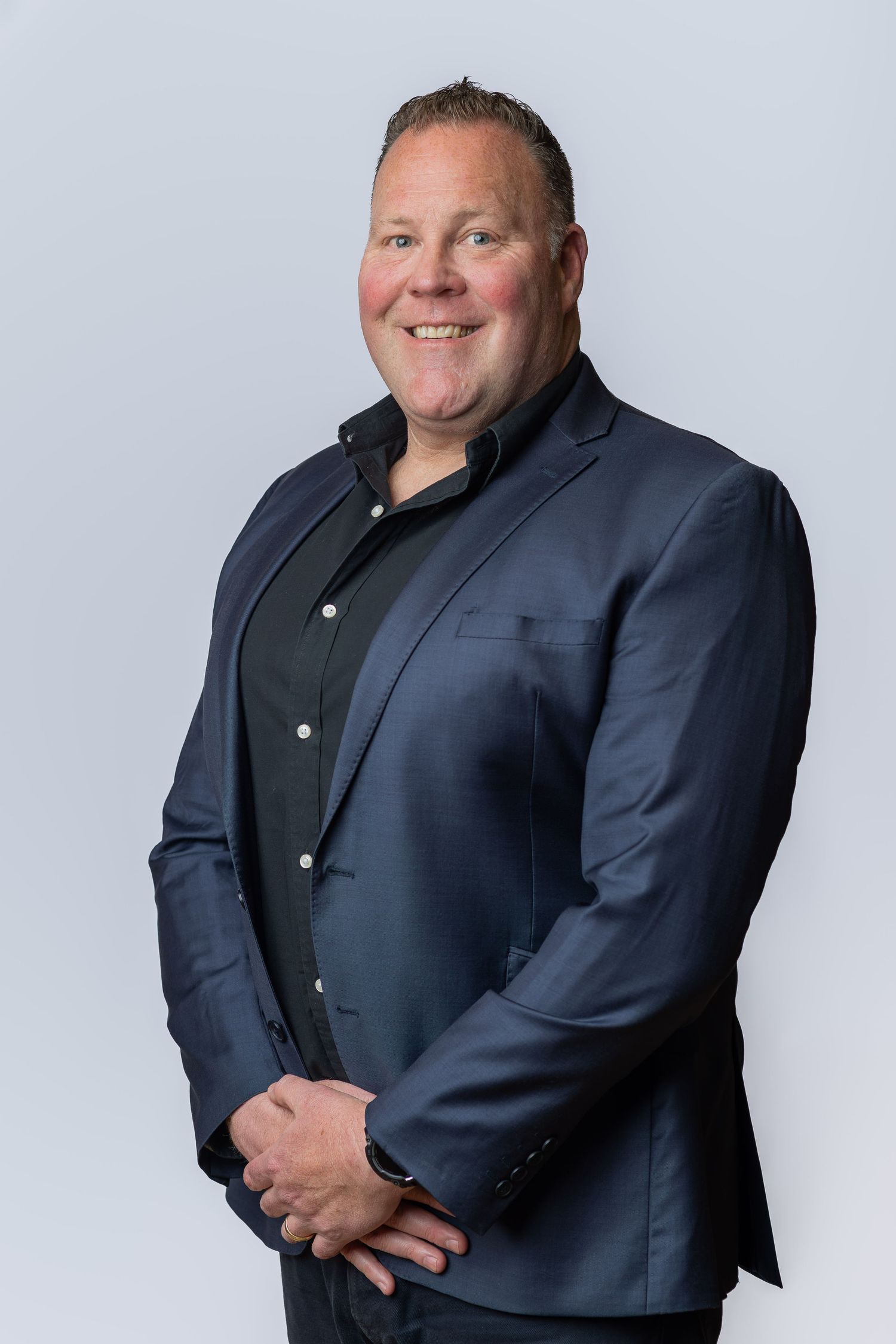 Professional man in navy blazer and black shirt smiling confidently against a light gray background.