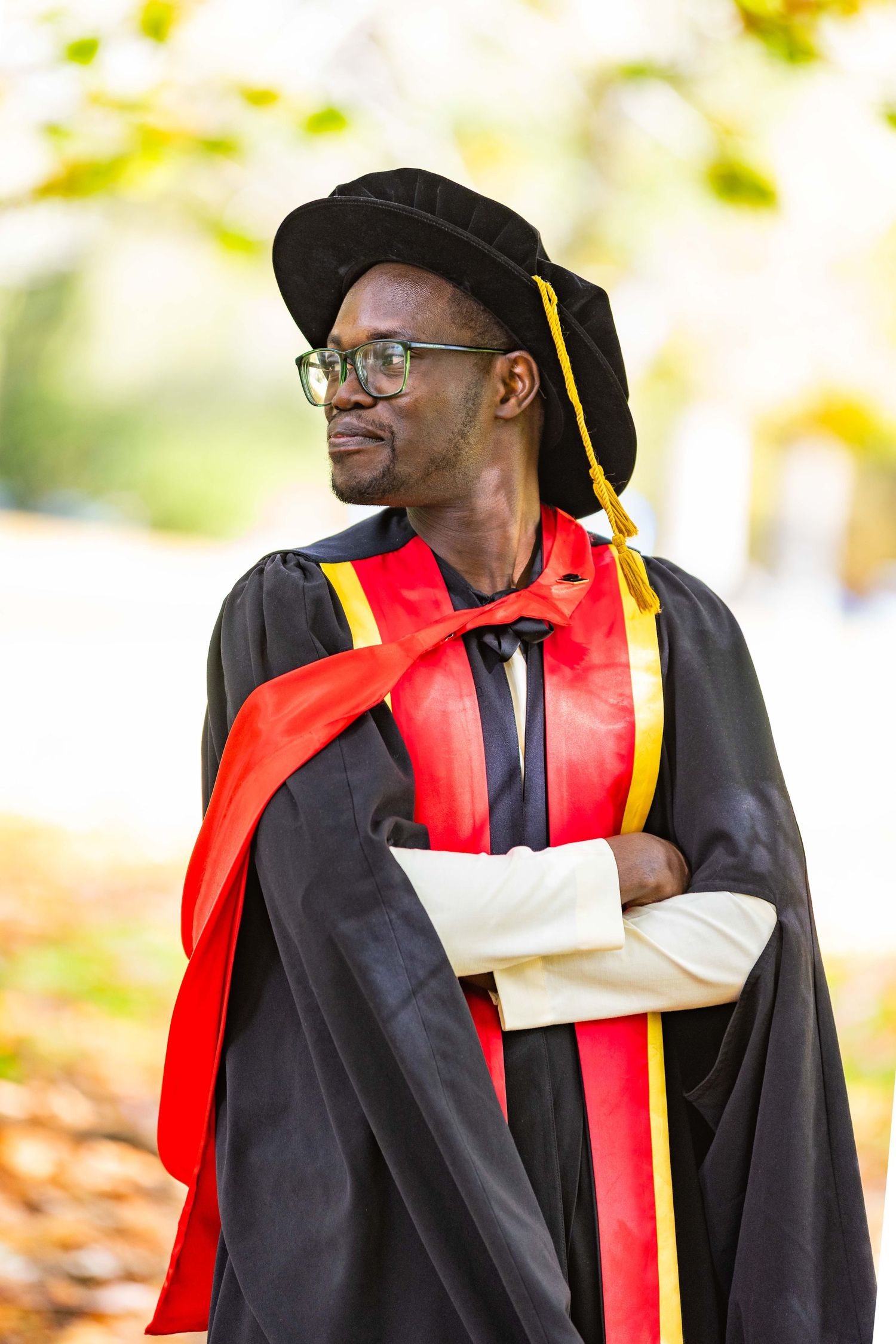 A proud graduate in black doctoral gown with red and gold trim poses with arms crossed outdoors.
