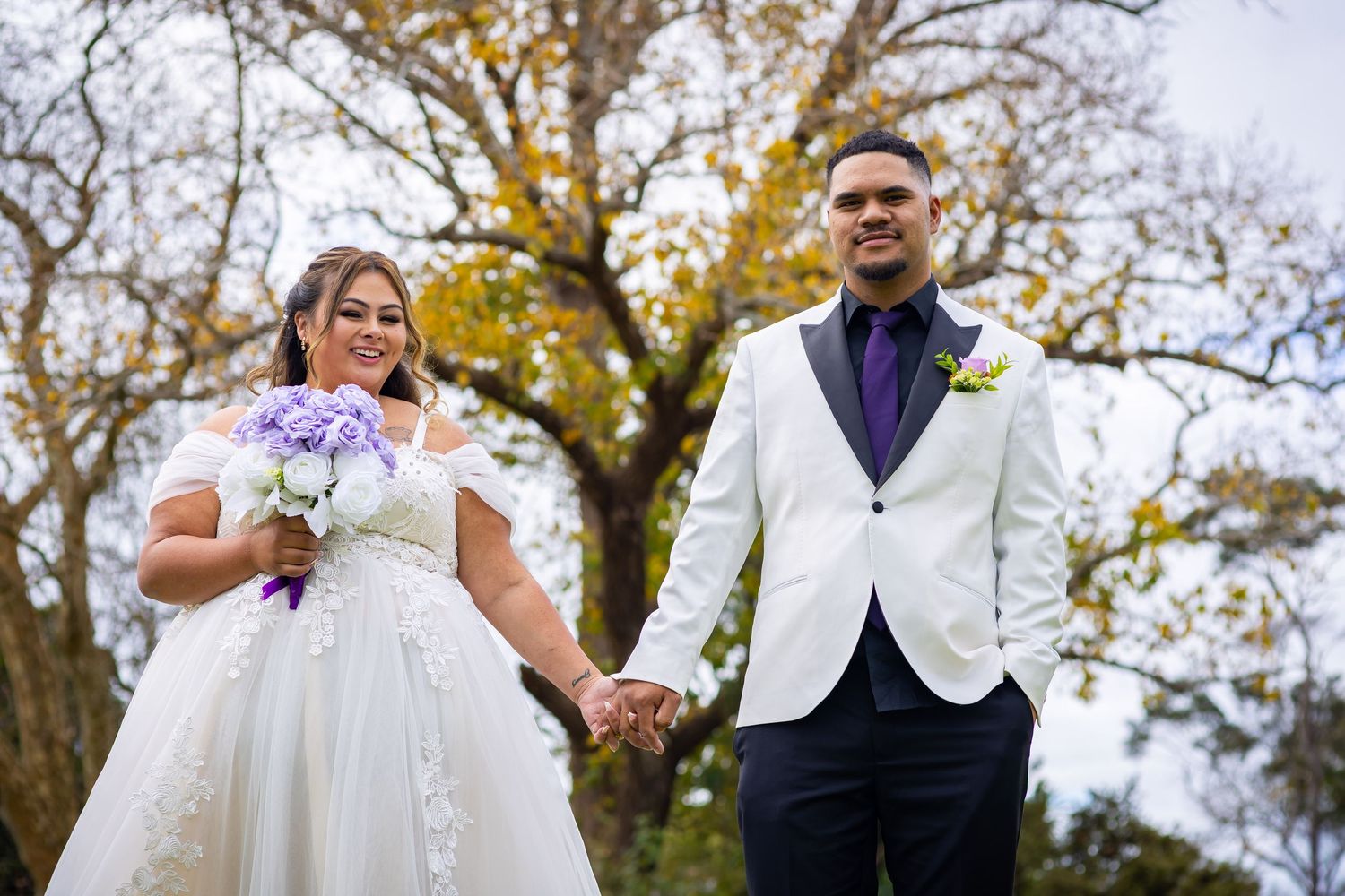 Bride and groom holding hands outdoors in wedding attire, she holds purple bouquet, autumn trees in background.