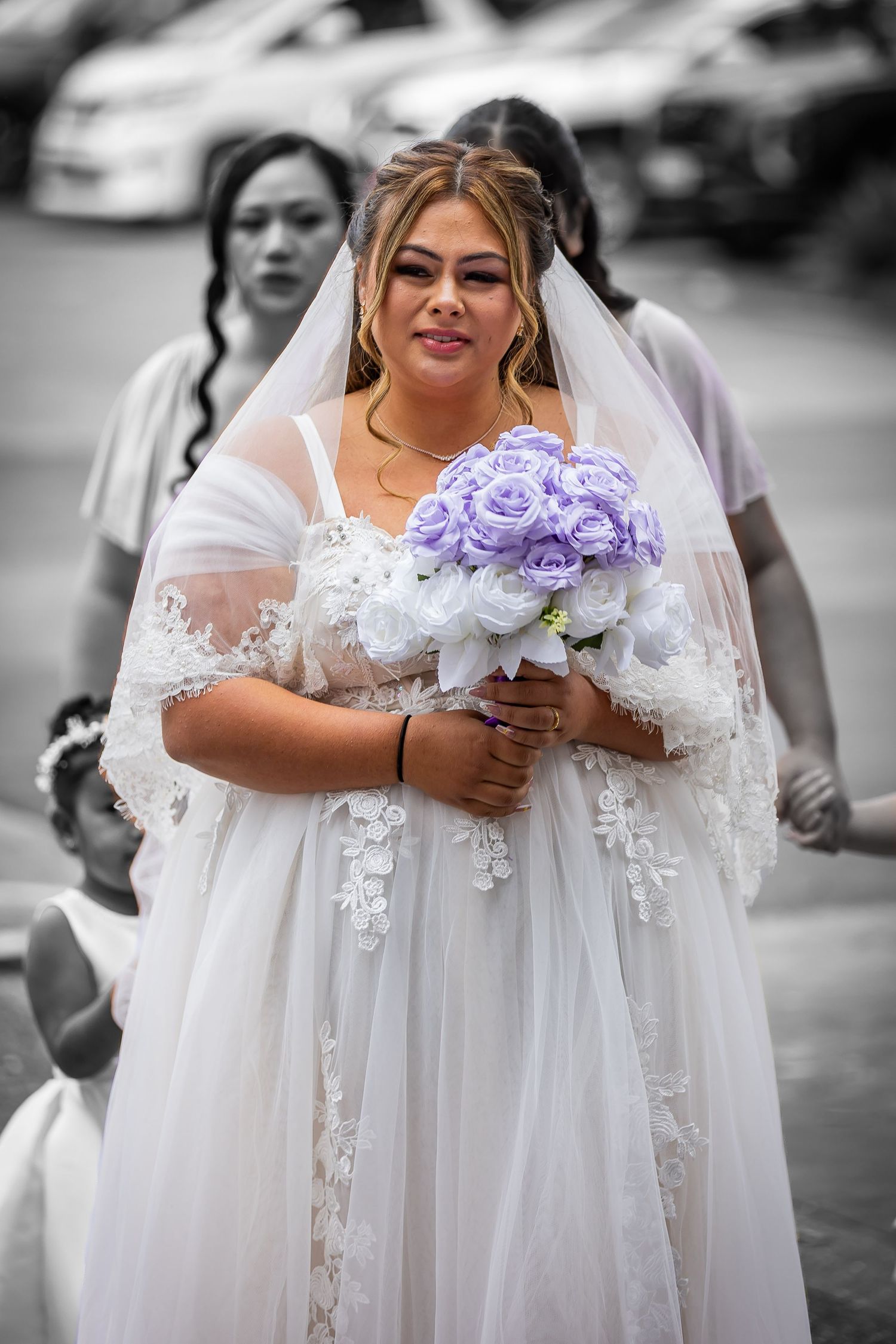 Bride in white lace gown and veil holds purple and white rose bouquet, with bridesmaids in background.