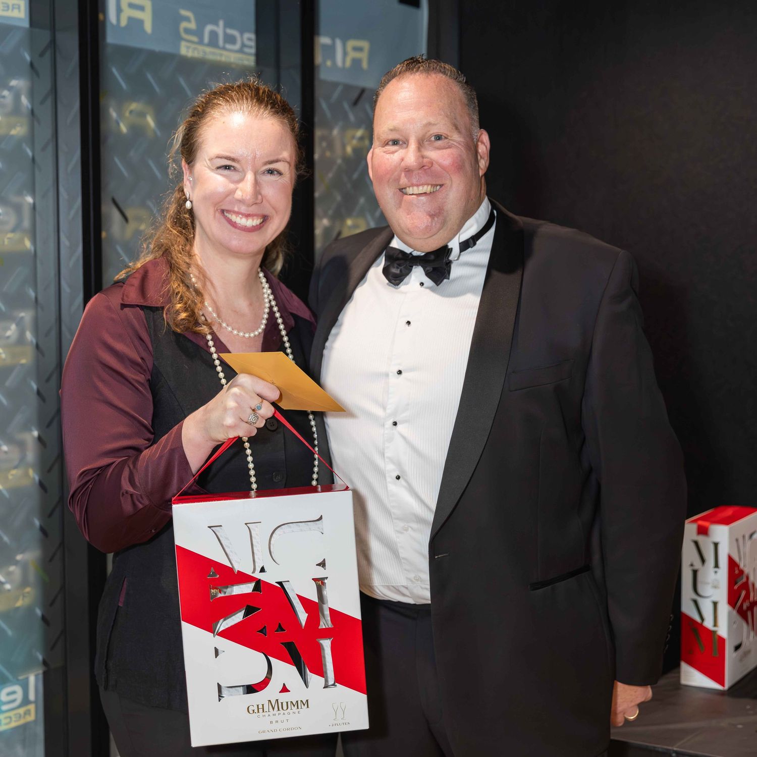 Man in tuxedo and woman in burgundy top smile at formal gala event, holding gift bag.