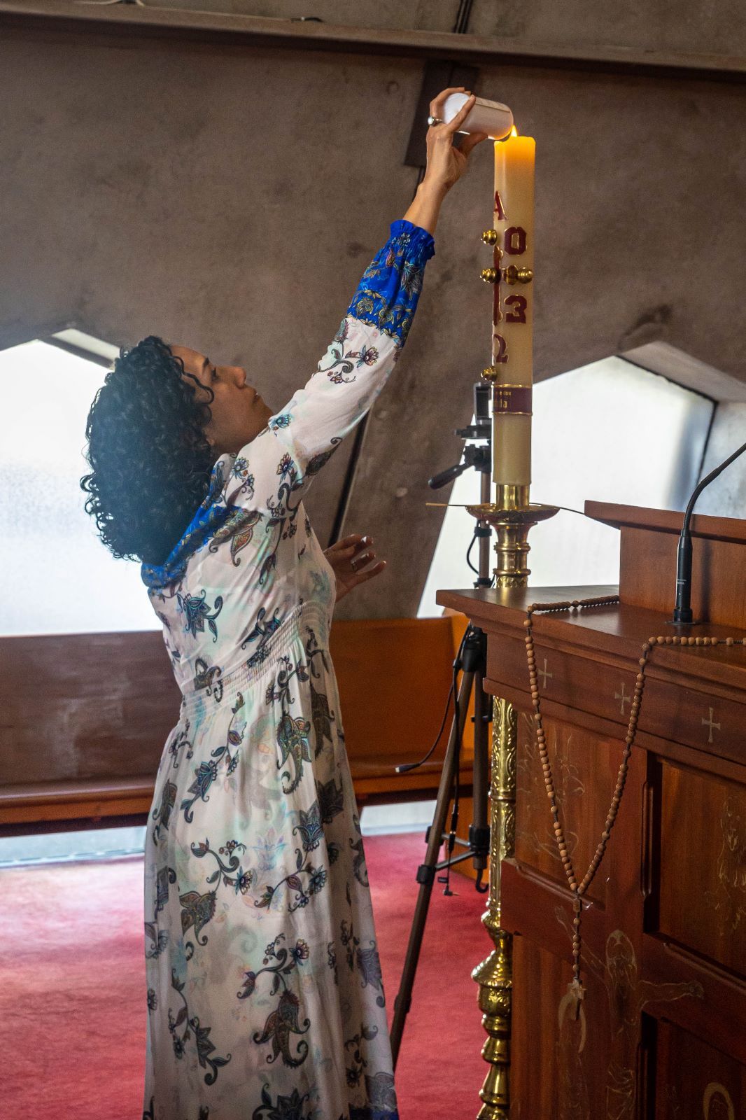 Woman in floral dress lighting a tall golden candle on an ornate candlestick in a church.