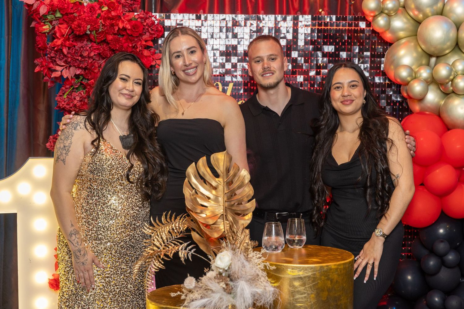 Four people posing together at a glamorous party with gold and red balloon decorations and a sequined backdrop.