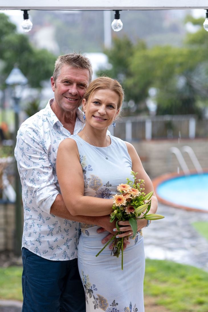 Middle-aged couple posing outdoors near a pool, woman holding a bouquet of flowers, both smiling warmly.