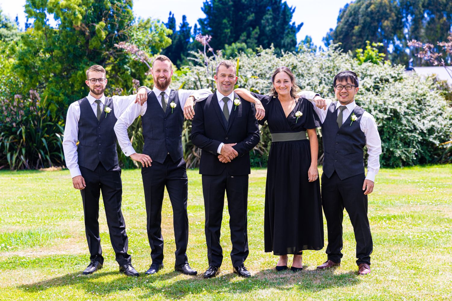 Wedding party of five posing outdoors on green lawn, wearing formal black attire with vests and boutonnieres.