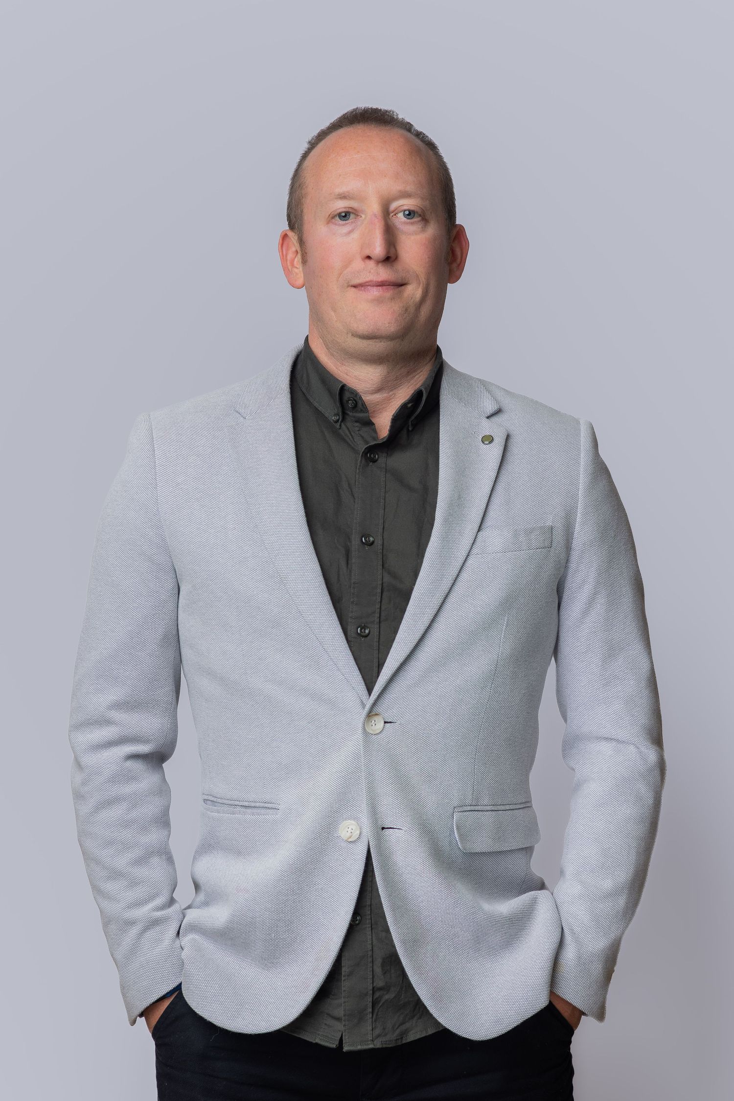 A professional man in a light gray blazer and dark shirt stands confidently with hands in pockets against a gray background.