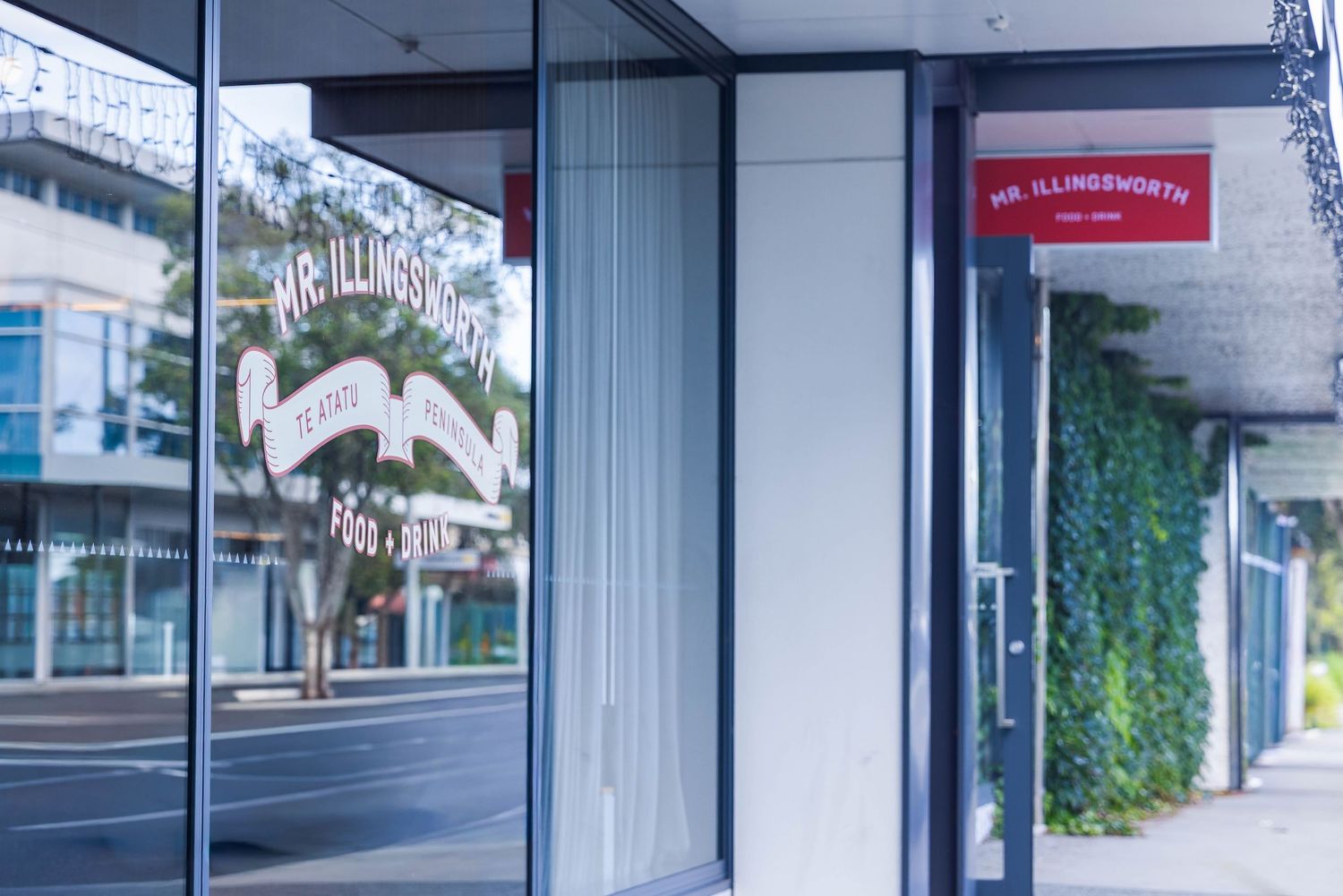 A storefront with glass windows and signage along a modern urban street with ivy-covered walls.