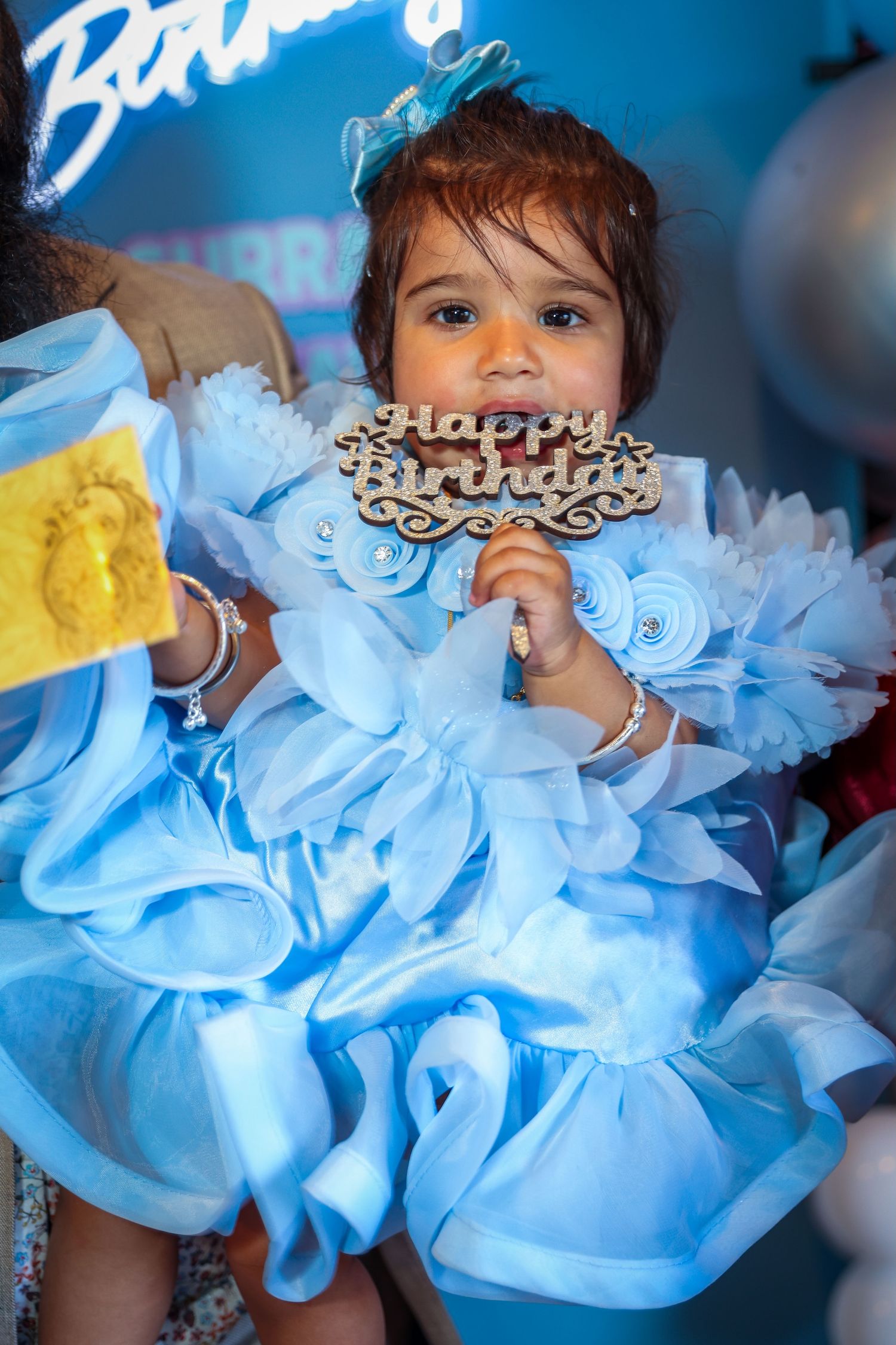 A baby girl in a blue ruffled dress holds a wooden Happy Birthday sign at her first birthday party celebration.