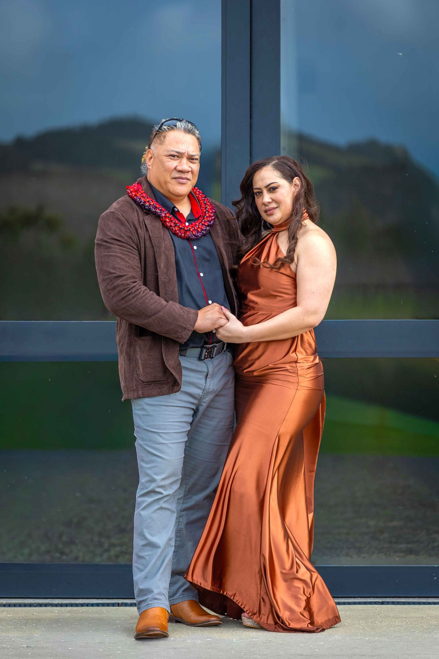Couple posing elegantly, woman in copper satin gown, man in brown jacket with red lei, standing before glass windows.