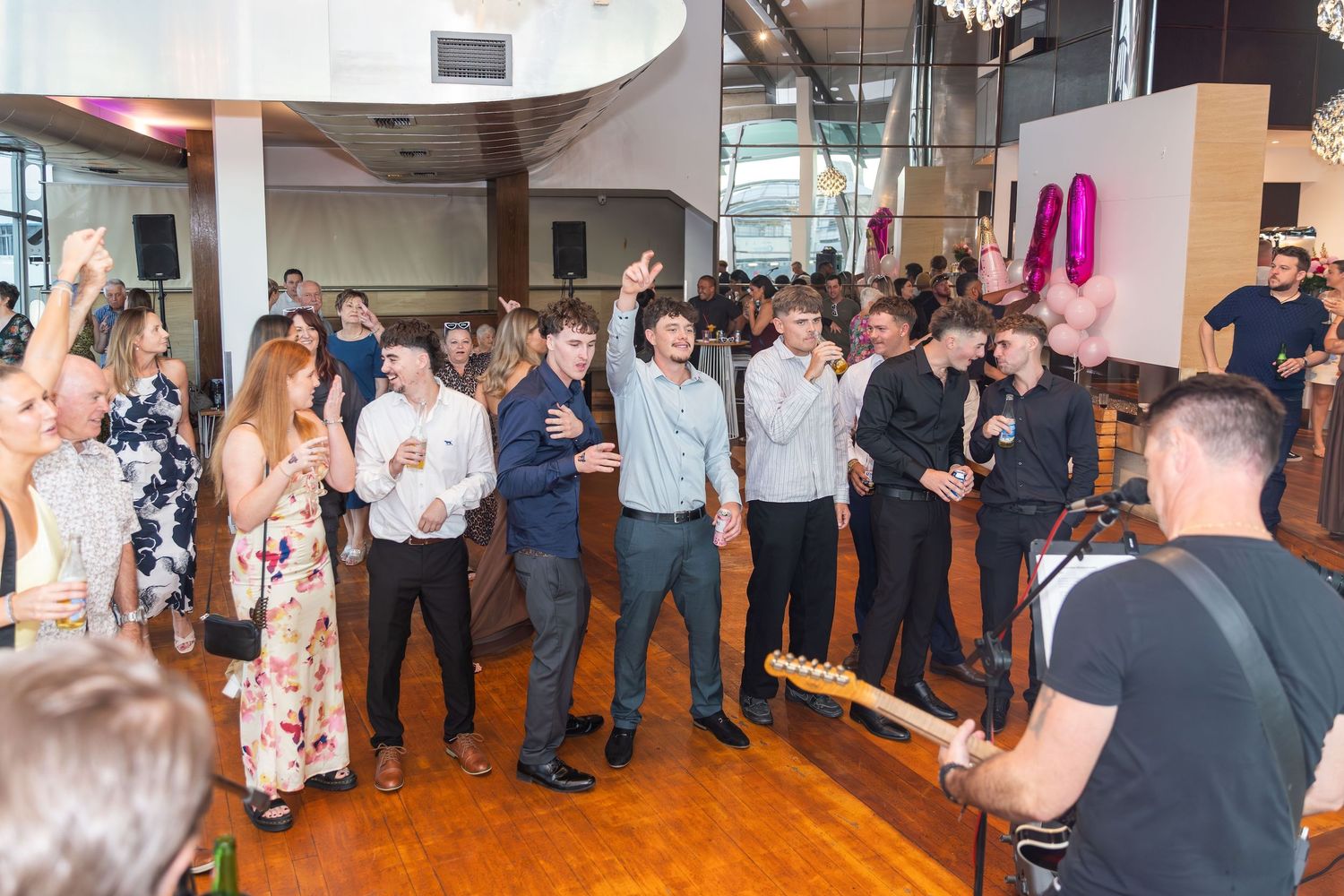 A lively indoor party with guests dancing and socializing near a live guitarist on a wooden dance floor.