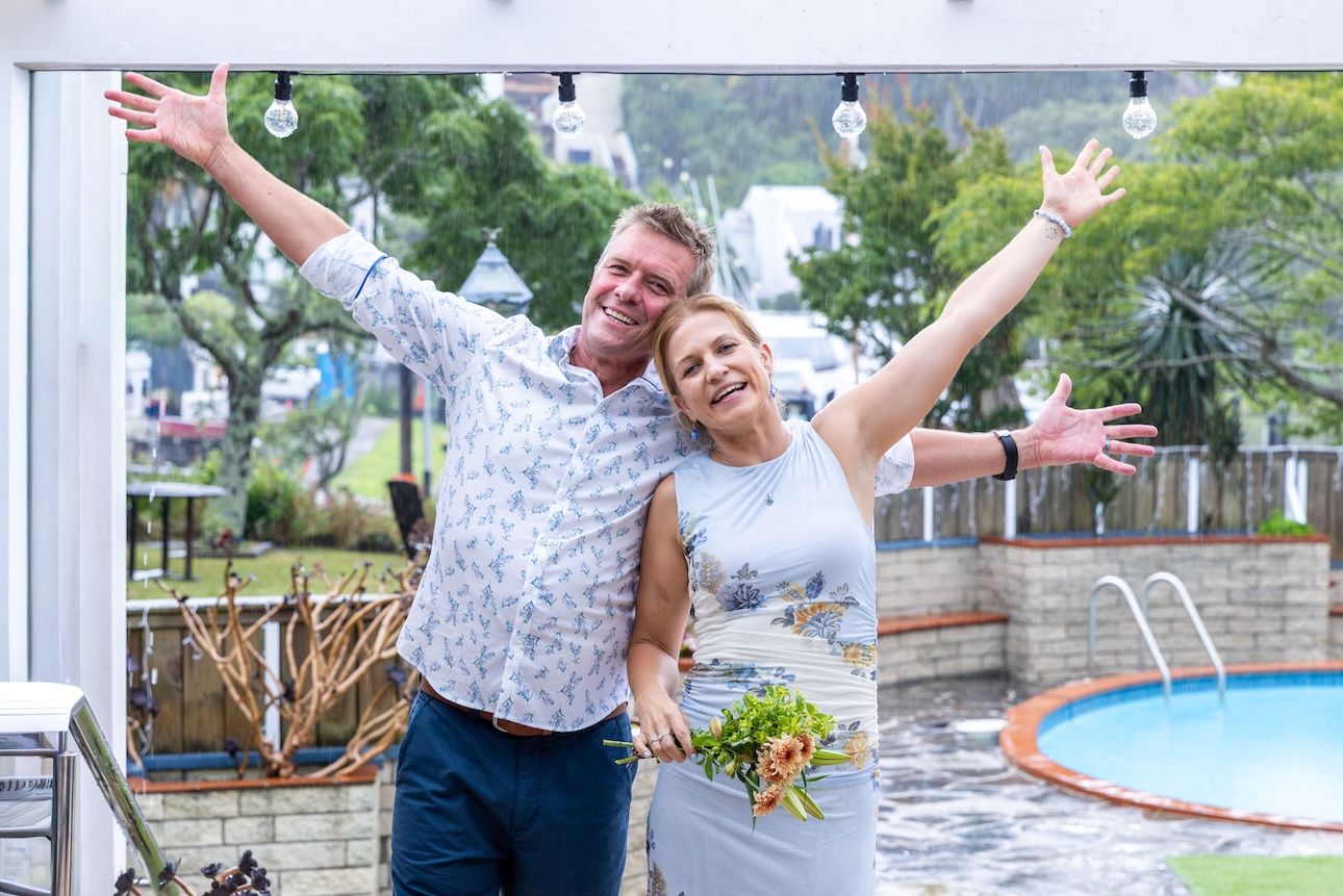 Joyful couple celebrating outdoors with arms raised near a pool, woman holding flowers, on a cloudy day.