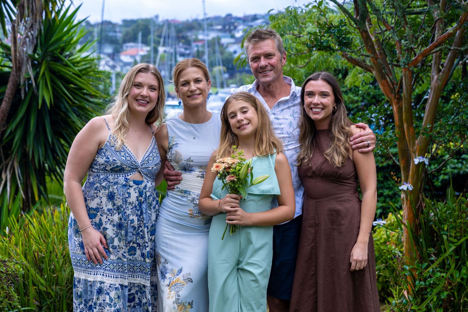 A group of people pose together outdoors in a lush tropical garden setting with palm trees and greenery in the background.