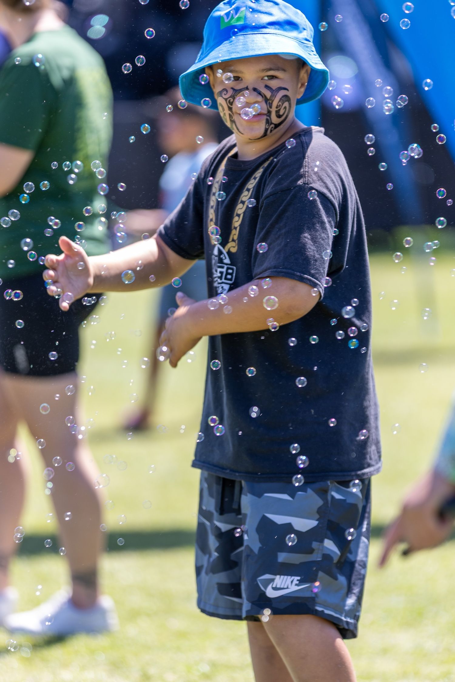 Children in blue hats play with bubbles on a grassy field during a sunny day.