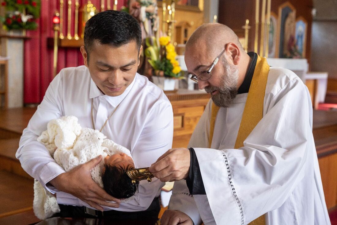 A priest performs a baptism ceremony on an infant held by a man in a white shirt inside a church.