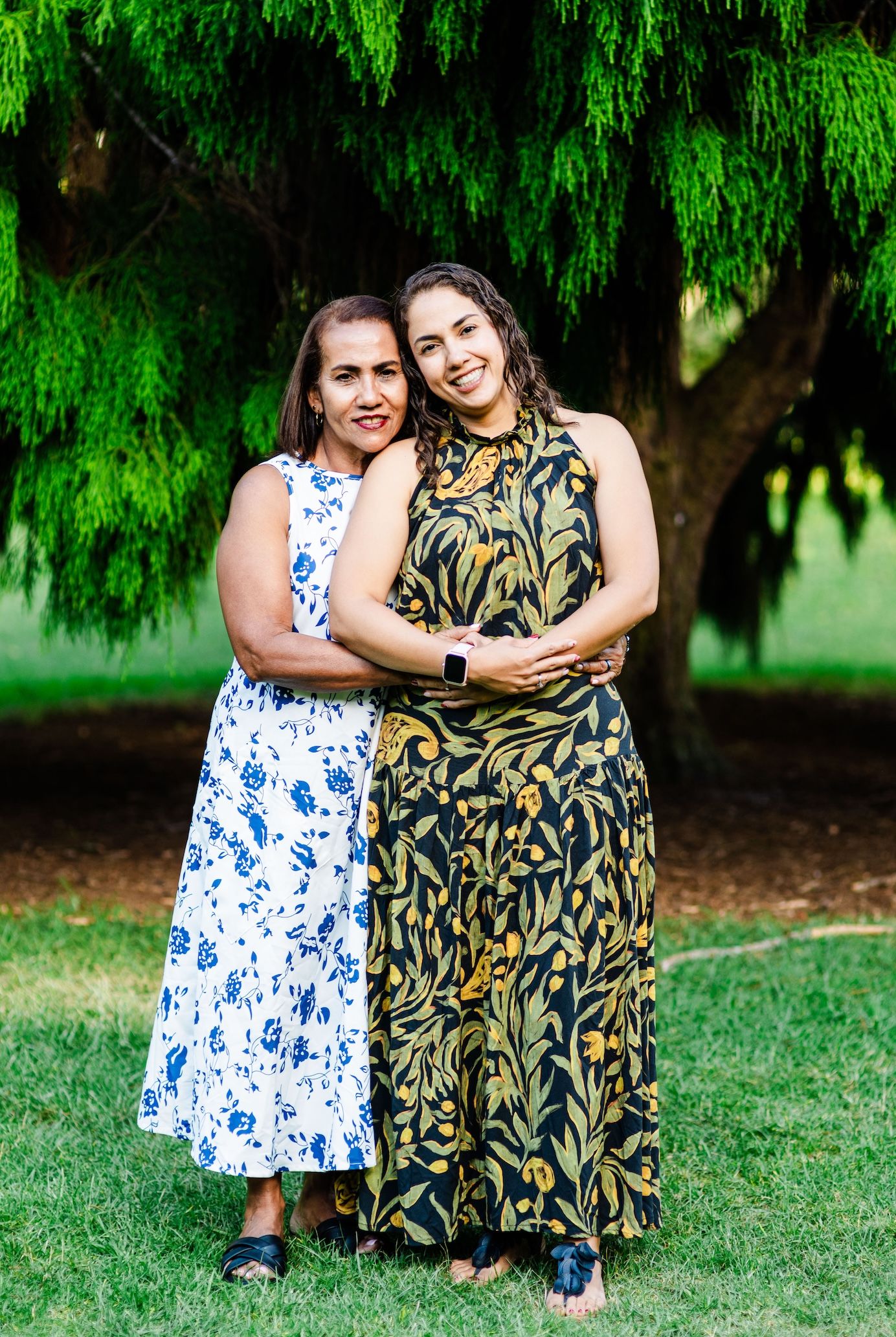 Two women in floral dresses embrace outdoors under a green tree on a sunny day.