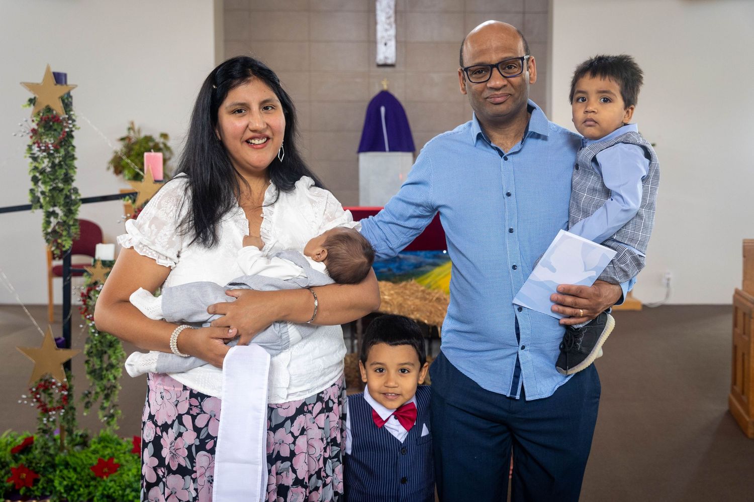 Family of four posing in a church, parents holding two young children, dressed in formal attire for a baptism ceremony.