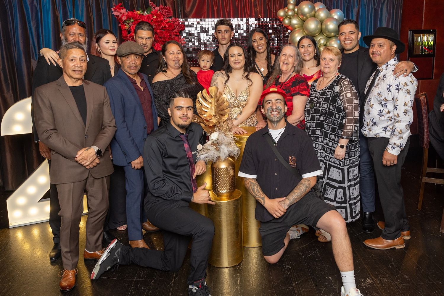 Large group of elegantly dressed people posing together at a festive party with balloons and decorative lighting.