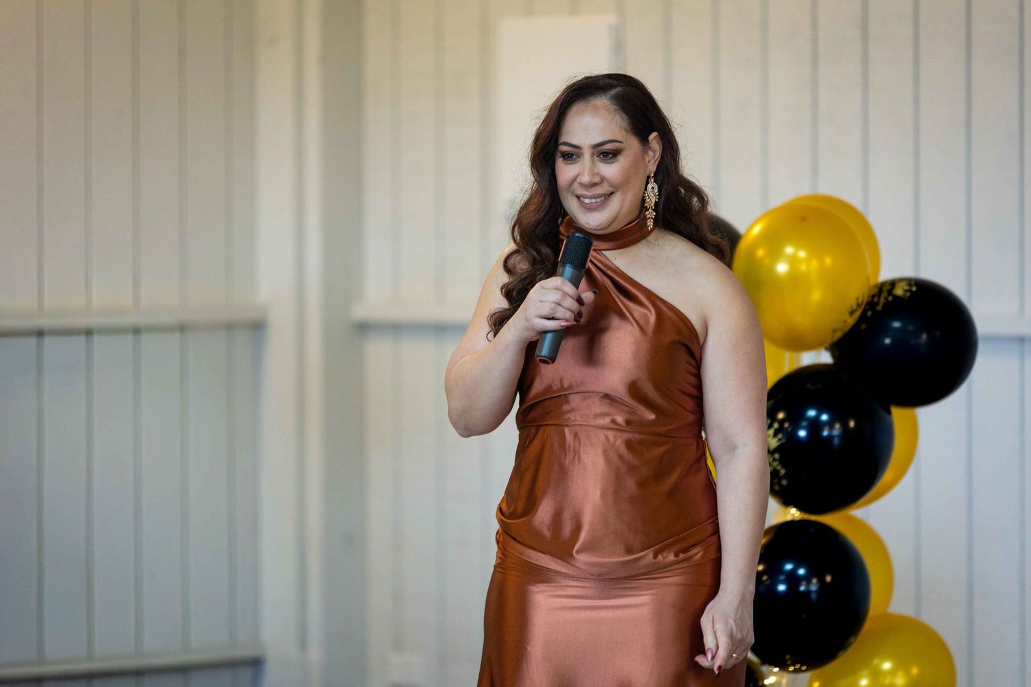 Woman in copper satin dress speaking into microphone at event with black and gold balloons.
