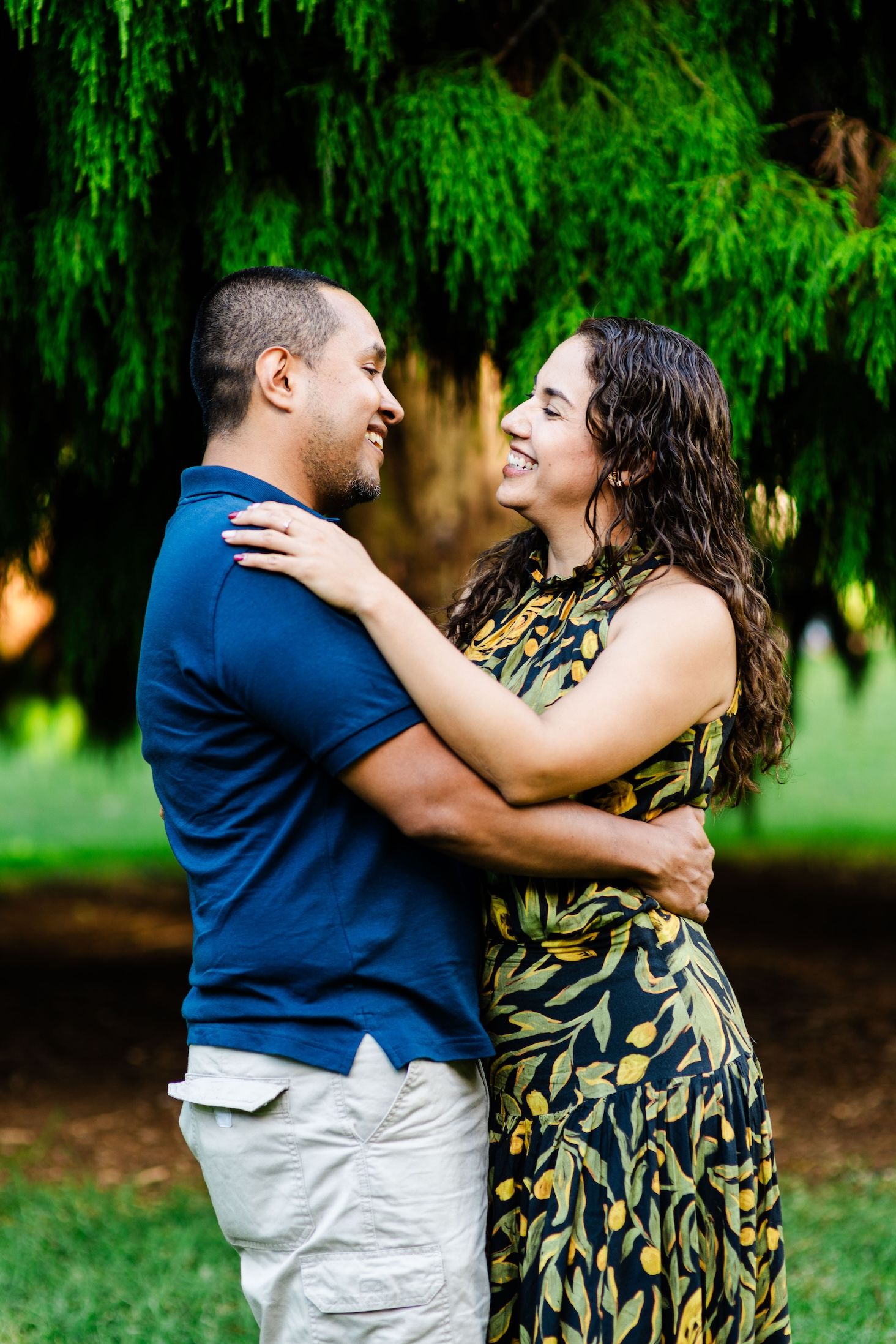 A couple shares a joyful embrace while dancing together in a park with lush green trees in the background.