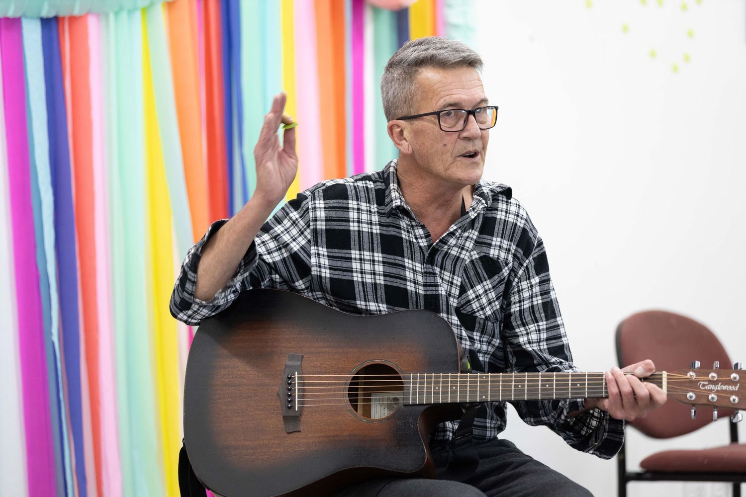 Man in plaid shirt playing acoustic guitar while gesturing with hand, colorful backdrop behind him.