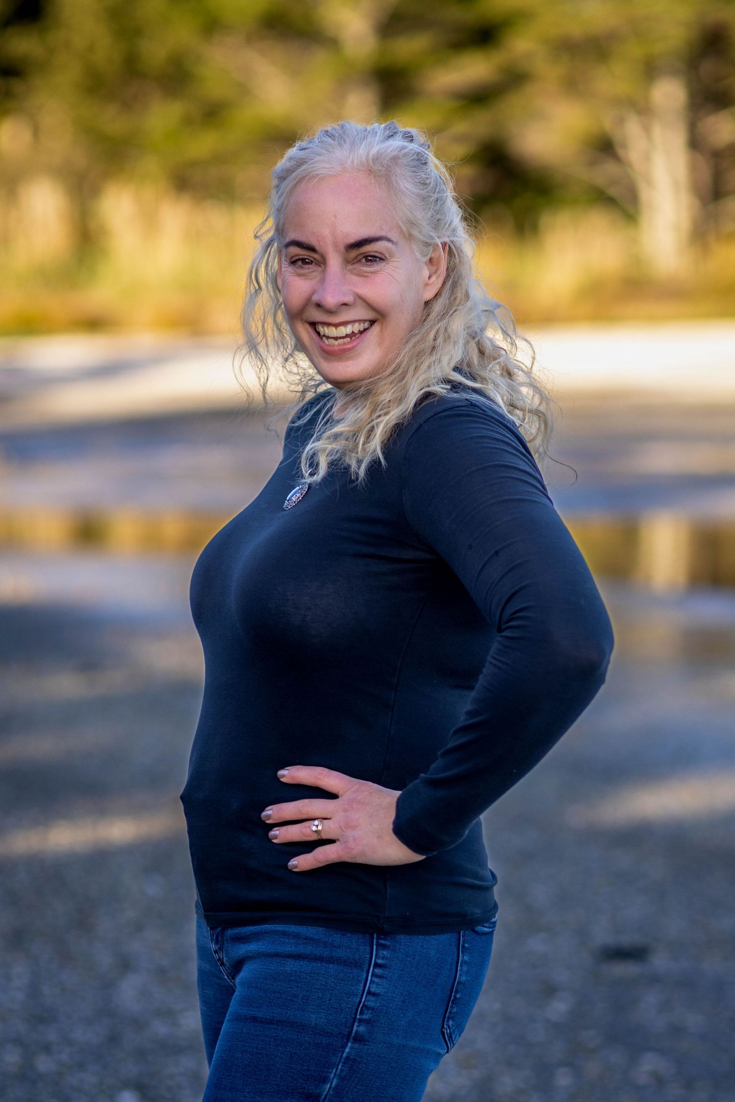 Smiling woman with silver hair in black turtleneck and jeans posing outdoors near trees.