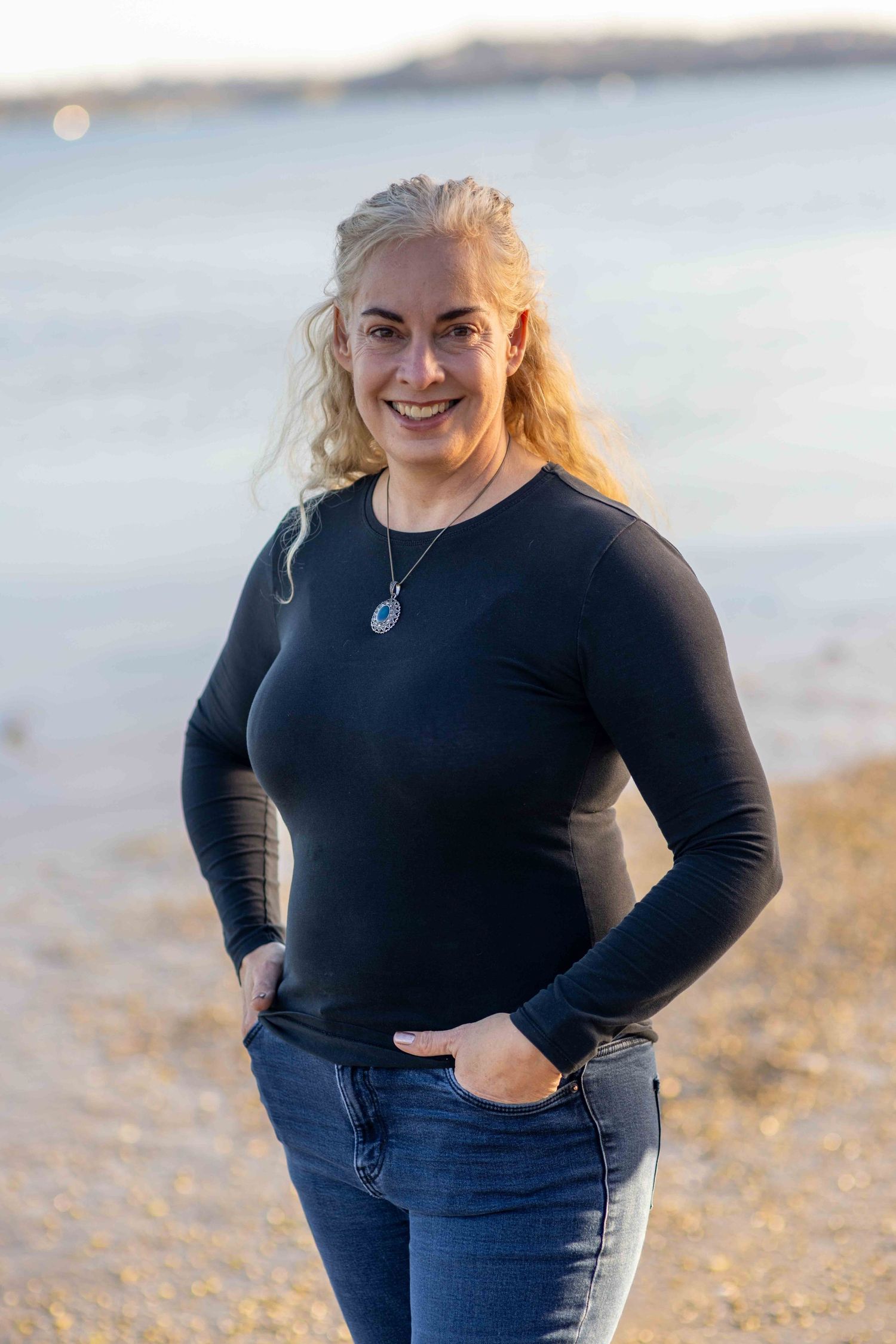Smiling blonde woman in black long-sleeve top and jeans standing on a sandy beach with water in the background.