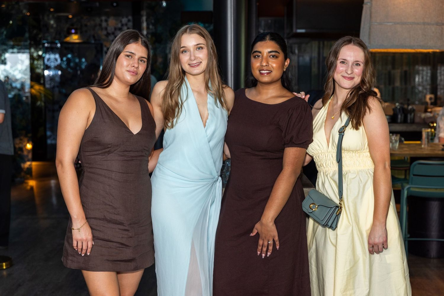 Four women in elegant dresses posing together at a formal event in a stylish venue.