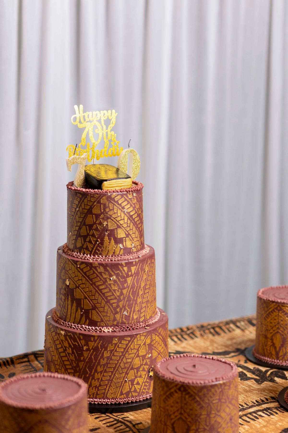 Three-tiered brown cake with intricate geometric patterns and a gold 'Happy Birthday' topper on a wooden table.