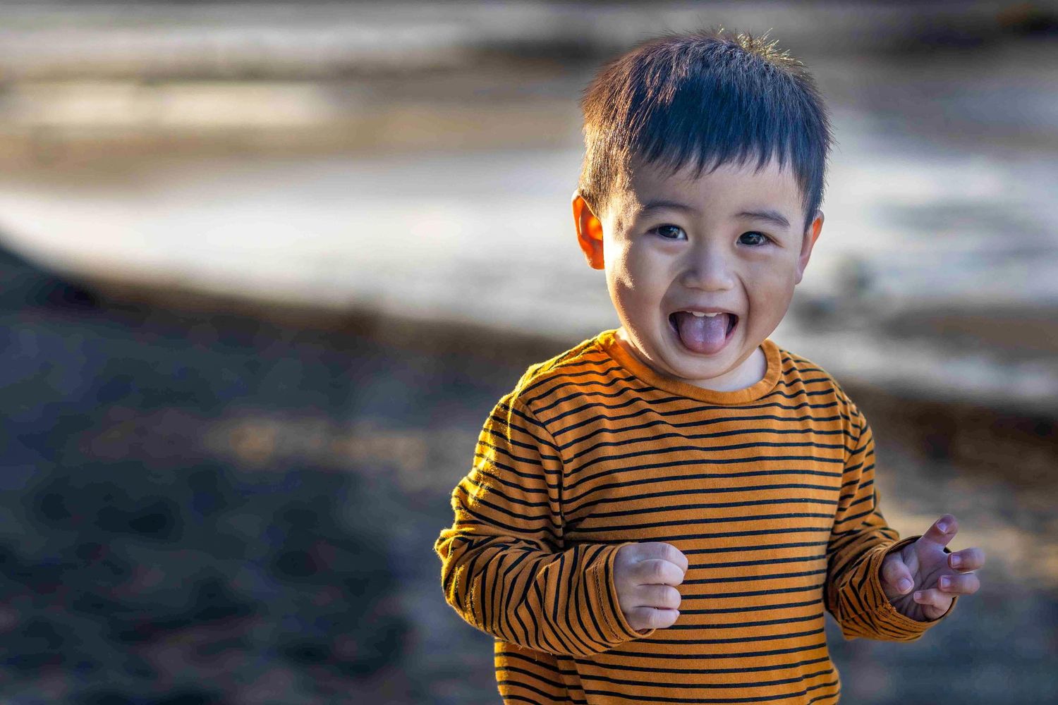 A joyful young Asian boy in a yellow striped shirt laughs outdoors near a rocky beach.