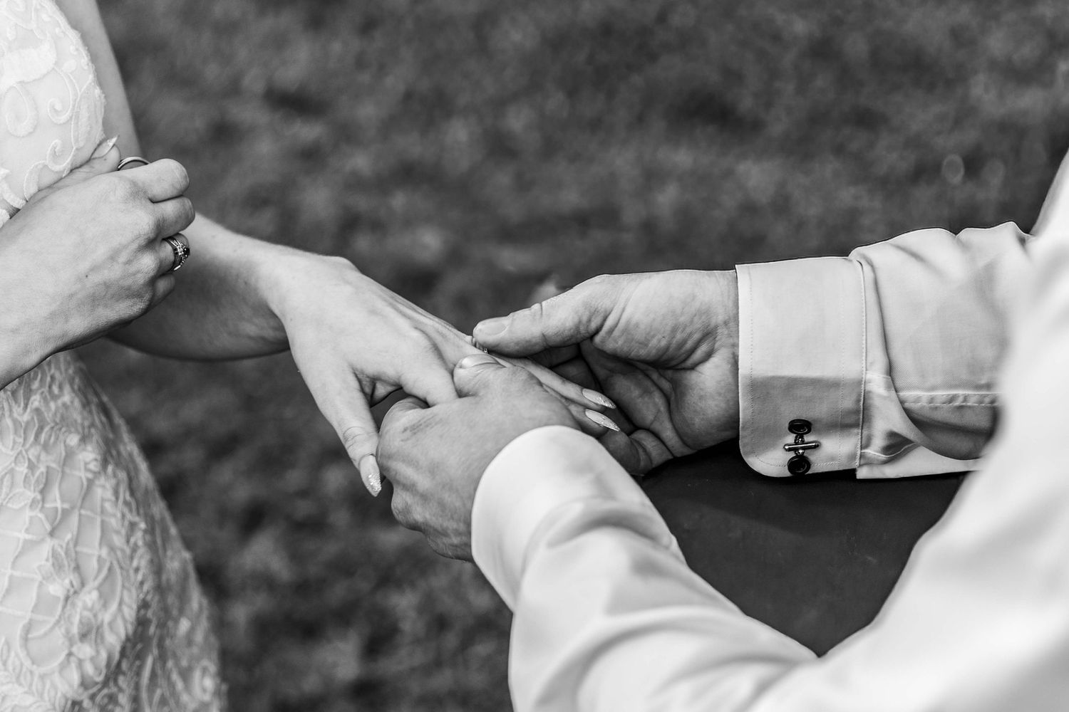 Black and white photo of groom placing wedding ring on bride's finger during outdoor ceremony.