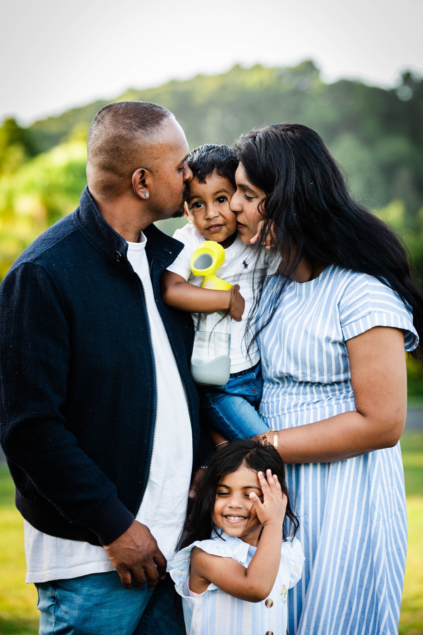 A loving family of four poses outdoors, parents kissing their toddler son while their smiling daughter stands in front.